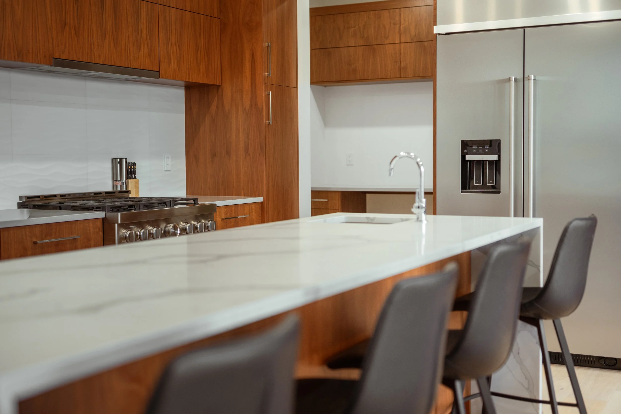 A modern kitchen with wooden cabinets, white countertops, a stainless steel stove, and a large refrigerator with an ice and water dispenser. There are chairs around the island and a sink in the foreground.