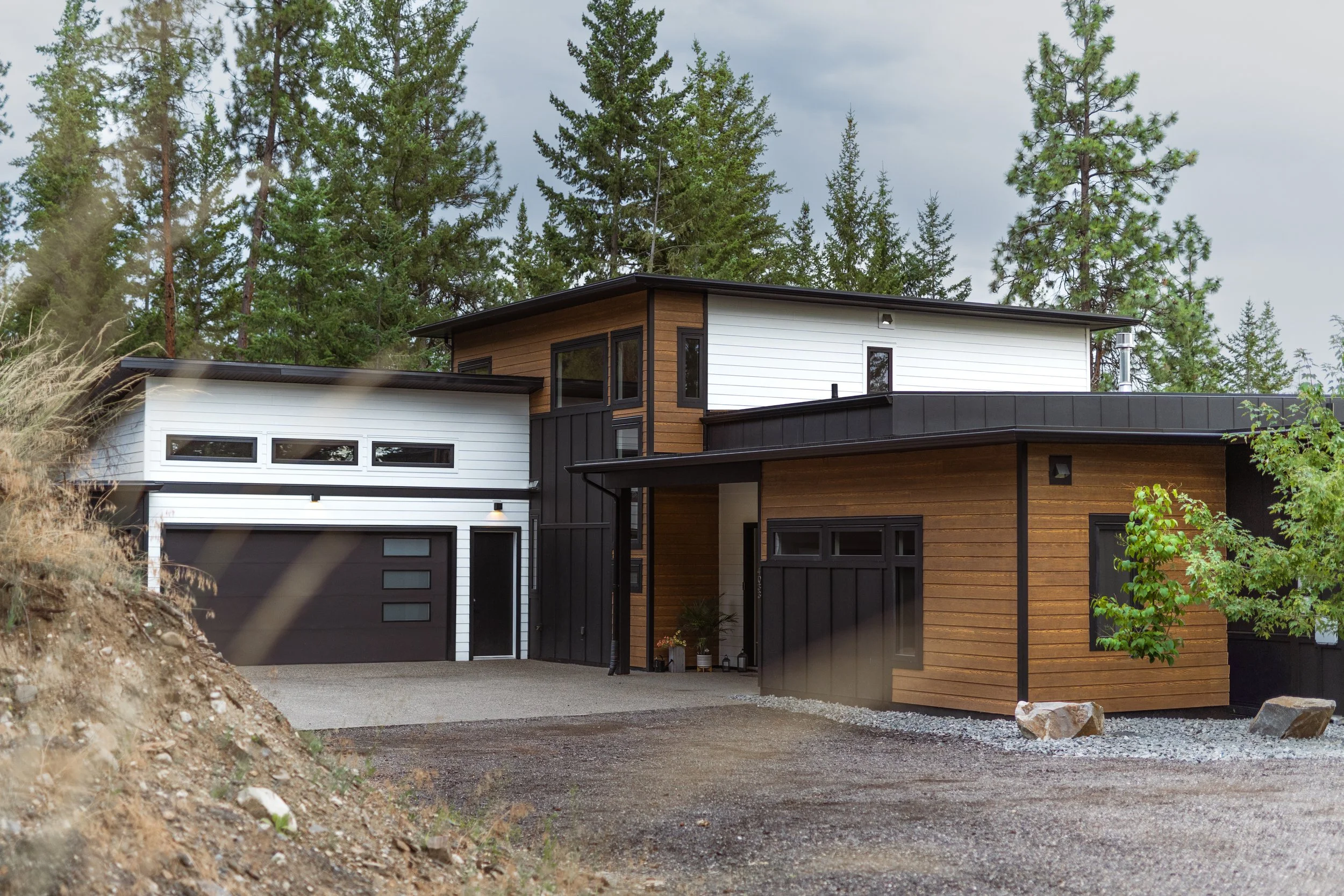 Modern two-story house with a flat roof, black and white exterior, and wooden accents, surrounded by tall pine trees and a cloudy sky.