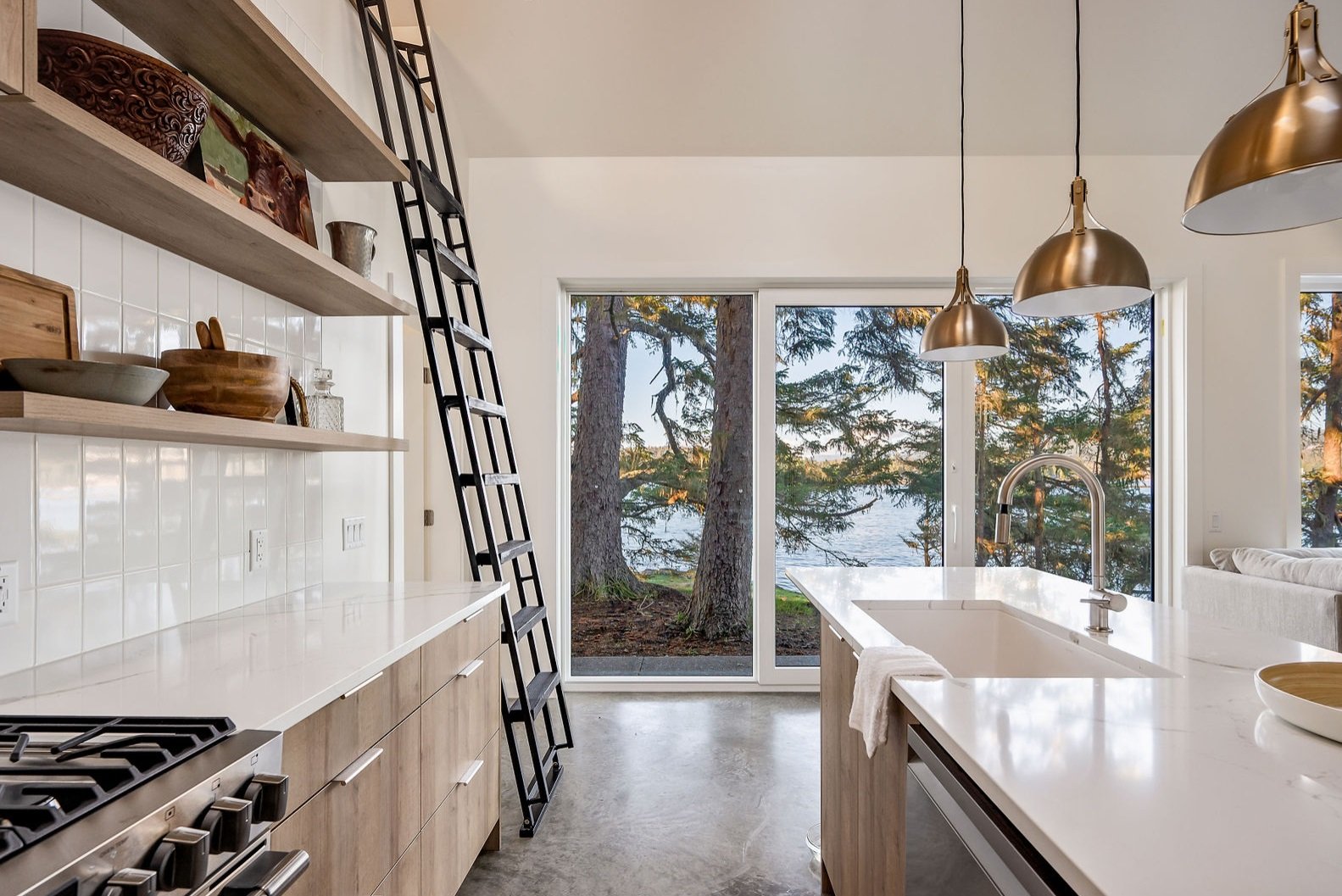Modern kitchen with white countertops, wooden cabinets, open shelves, a gas stove, large windows, and a view of trees and water outside.