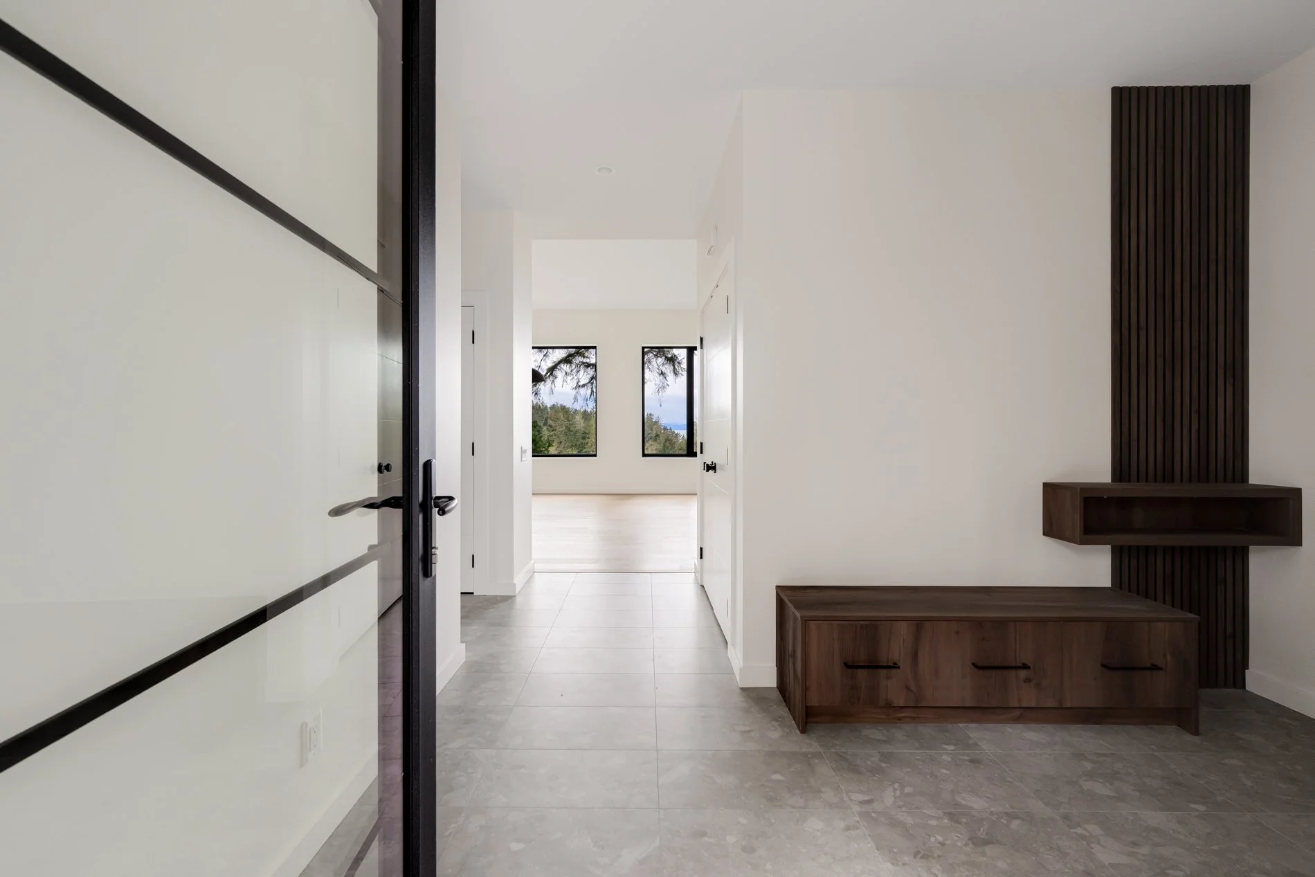 Modern minimalist home entryway with white walls, light-colored tile floor, and built-in wooden bench with open storage. Large windows in the background offer a view of greenery and trees.