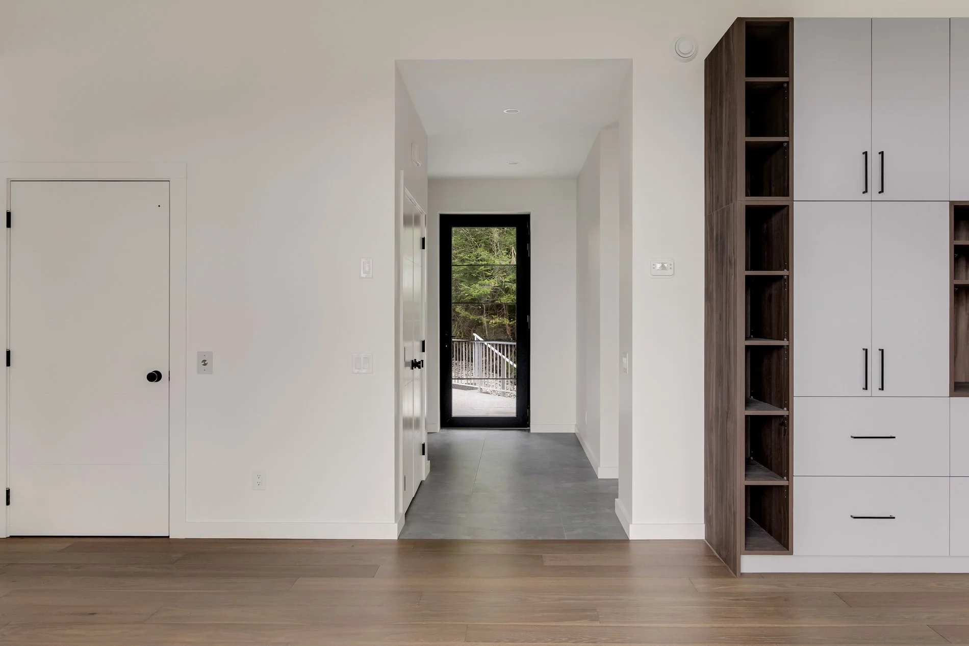 Interior of a modern home with white walls, a doorway leading outside to a deck with trees, and a built-in bookshelf with dark wood accents.