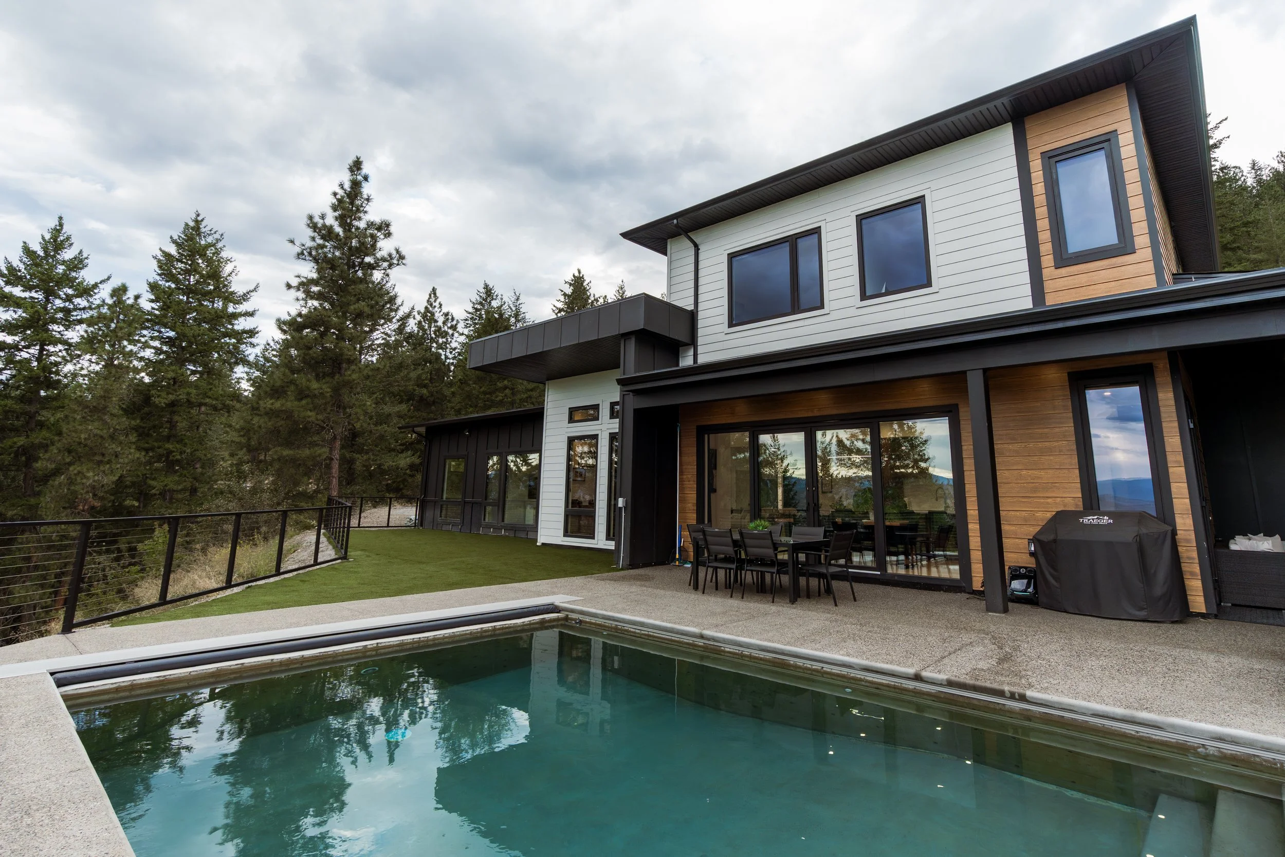 Modern house with large windows, surrounded by trees, with a backyard pool and patio area with outdoor furniture.