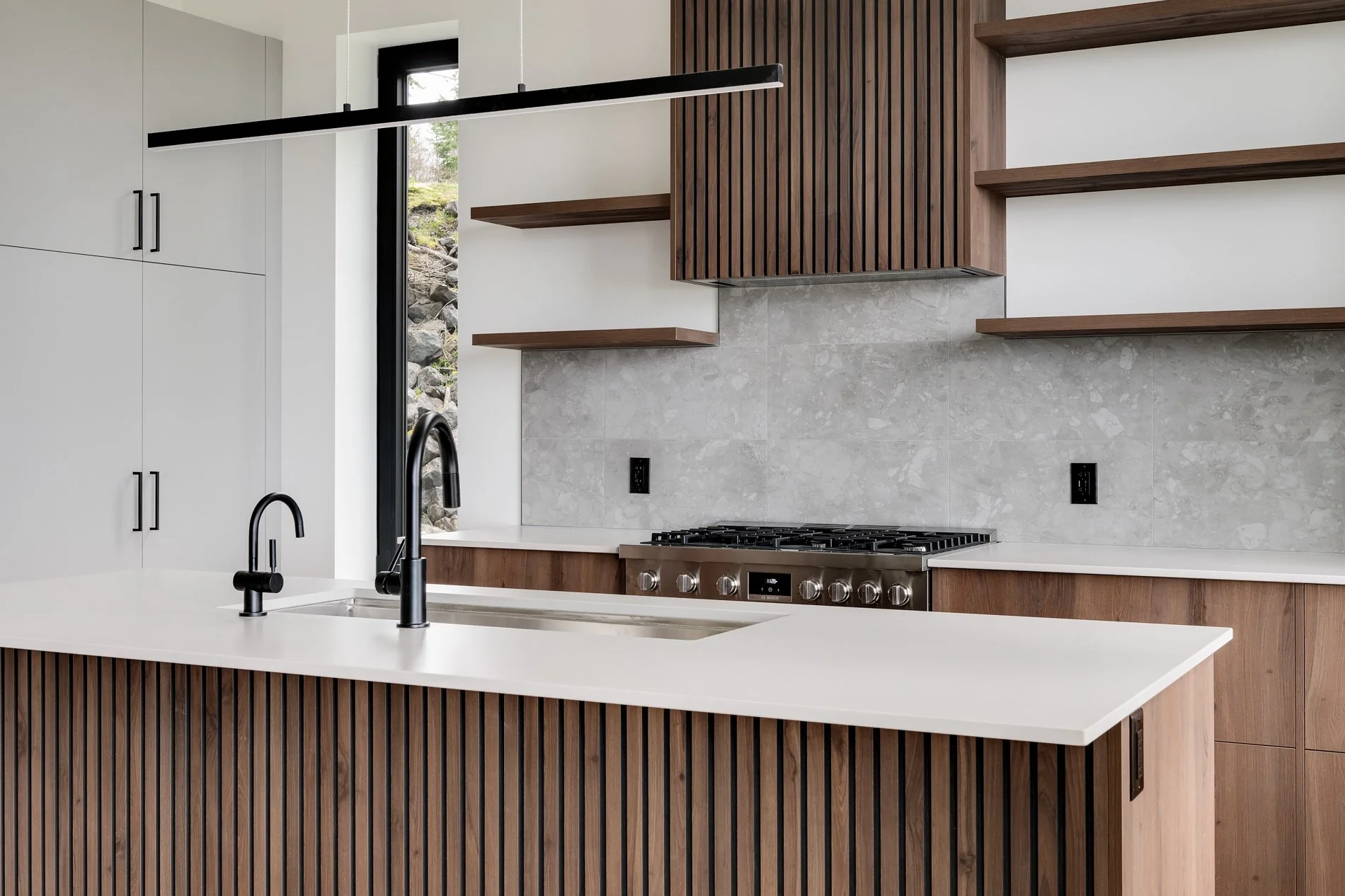 Modern kitchen with white cabinets, black fixtures, light gray marble backsplash, wooden open shelves, stainless steel gas range, and a white island with a black faucet.