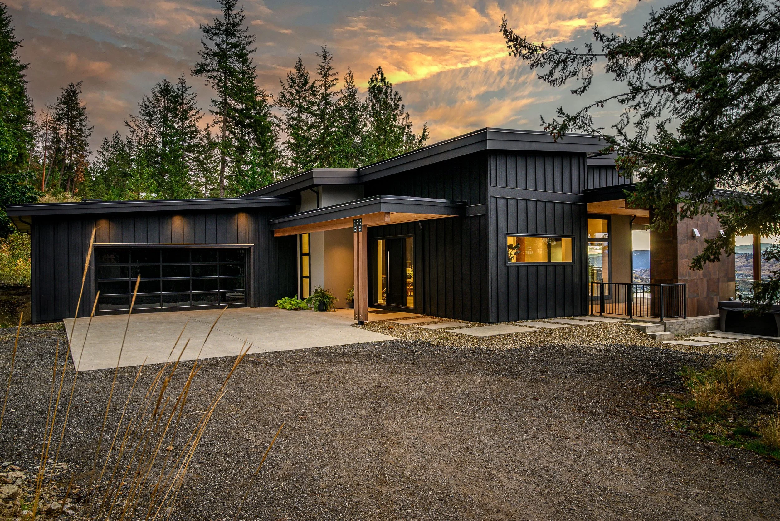 Modern black house with large windows, a garage with a black door, a concrete driveway, and a small front porch with wooden beams, set against a backdrop of trees and a sunset sky.