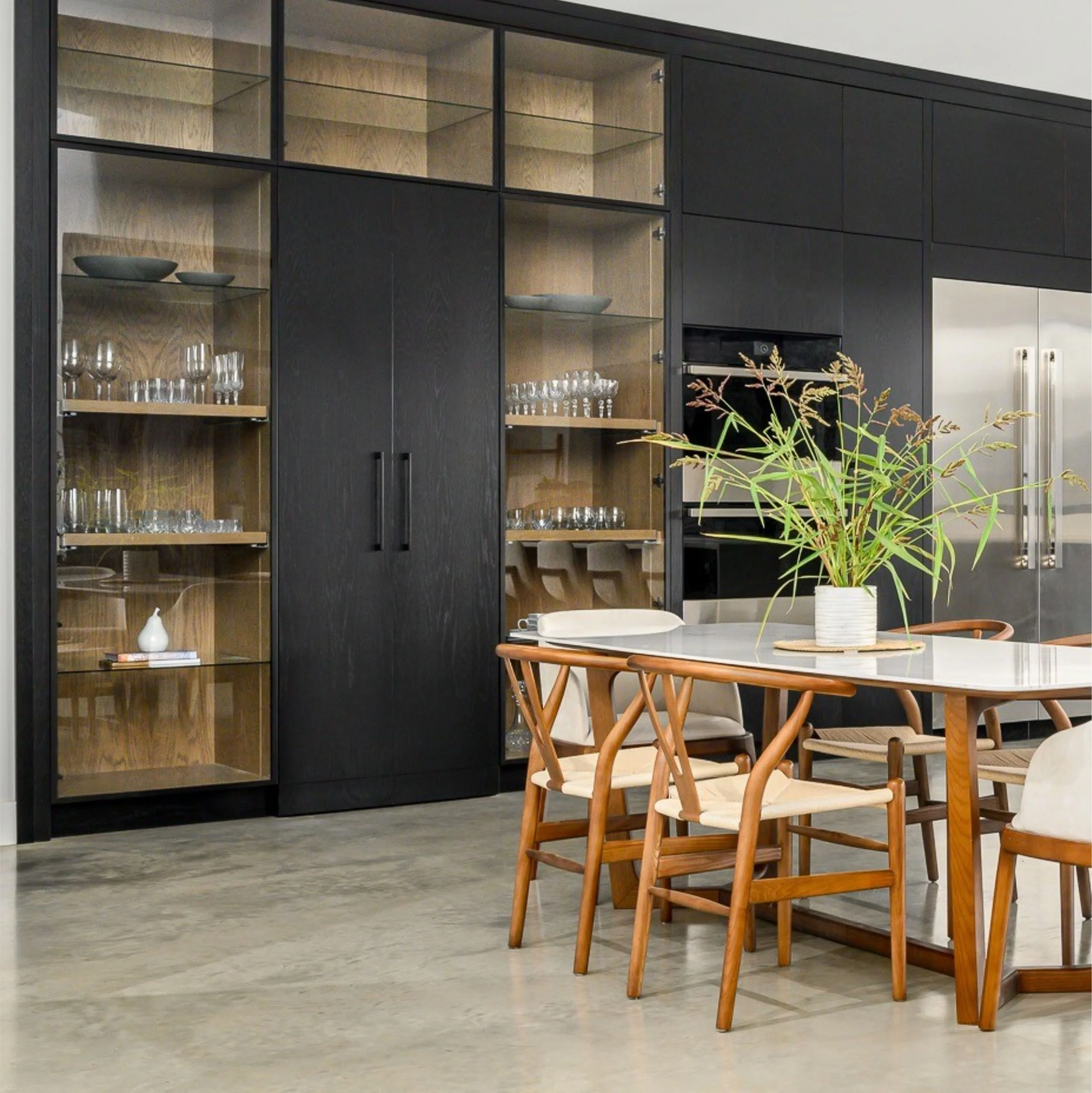 Modern dining area with wooden chairs around a table and a black cabinet with glass doors displaying glassware and decorative bowls.
