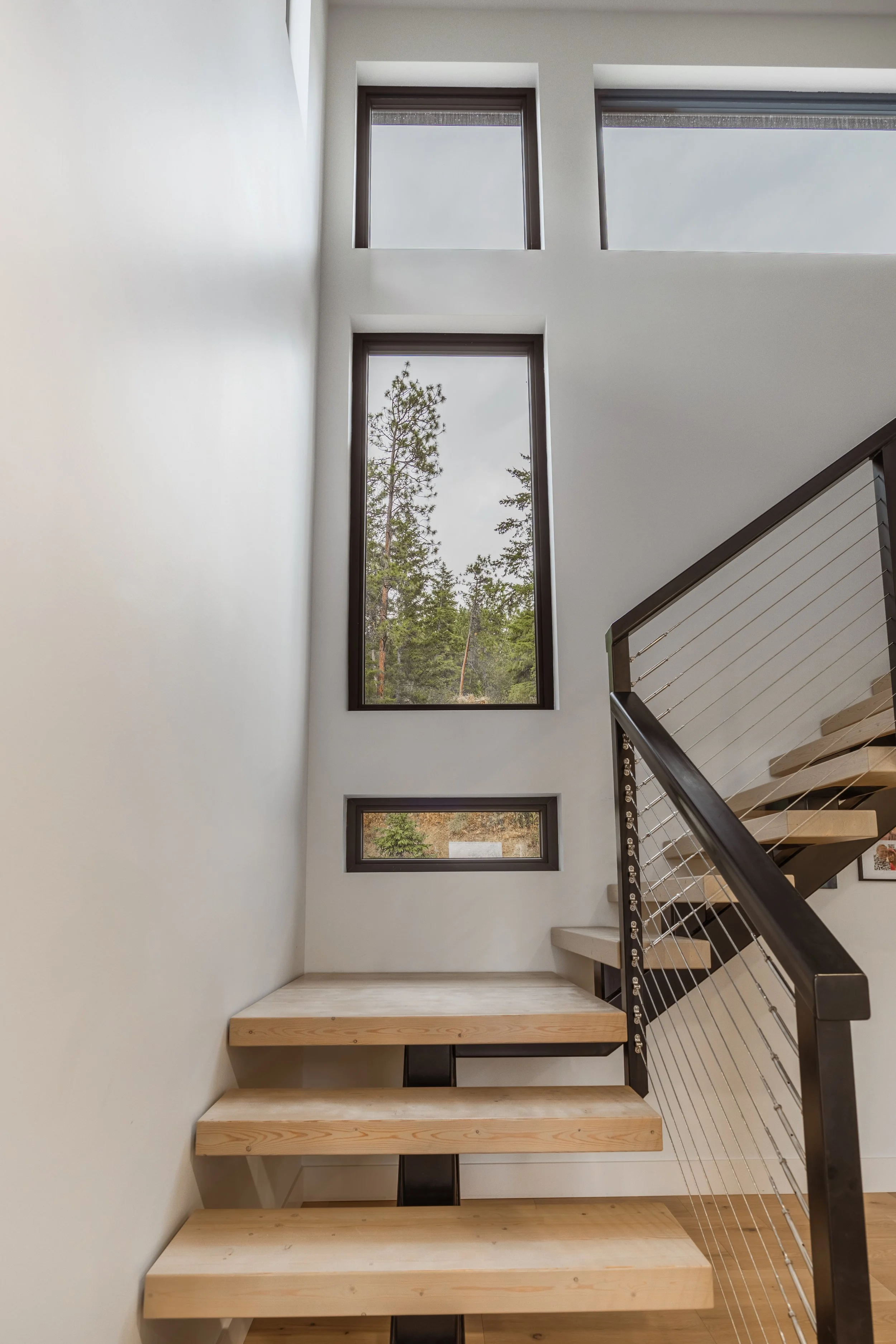 Interior view of a modern staircase with wooden steps, a metal and cable railing, and large rectangular windows looking out onto a forest landscape.