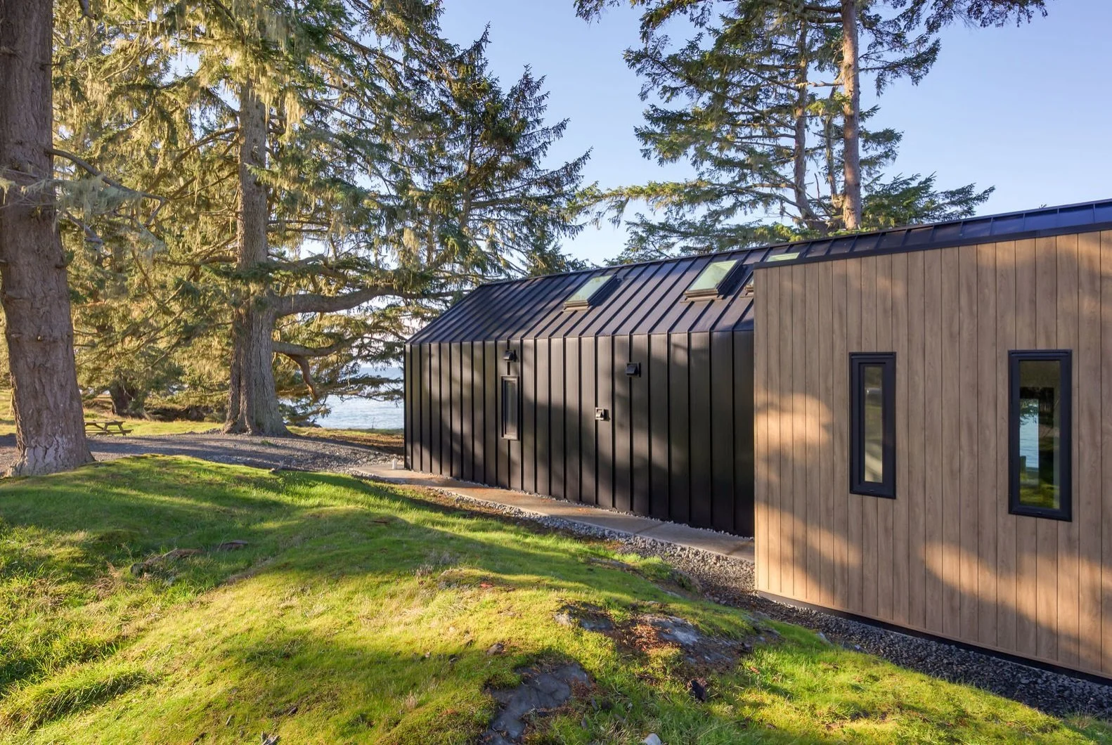 A modern house with black metal and wooden exterior walls, tall trees, and a grassy yard, near a body of water.