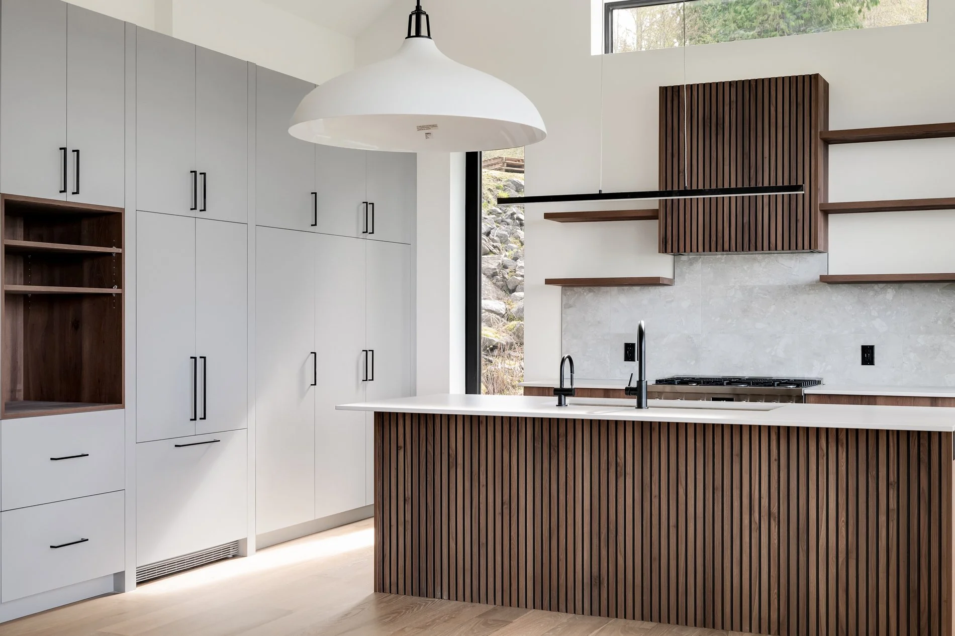 Modern kitchen with white cabinetry, dark wooden accents, a large white pendant light, and a window overlooking a rocky outdoor landscape.