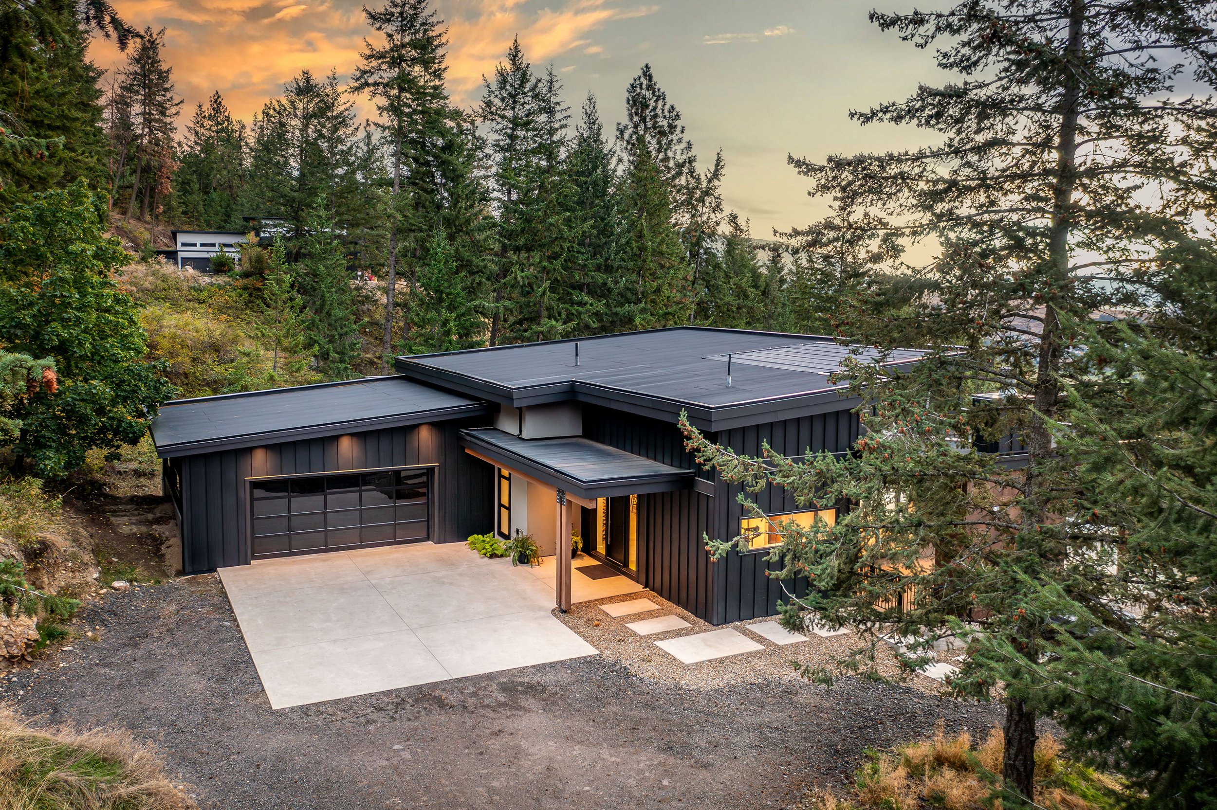 A modern house with a black exterior and flat roof, surrounded by tall pine trees, with a concrete driveway and a wooden pathway leading to the entrance, set in a wooded area during sunset.