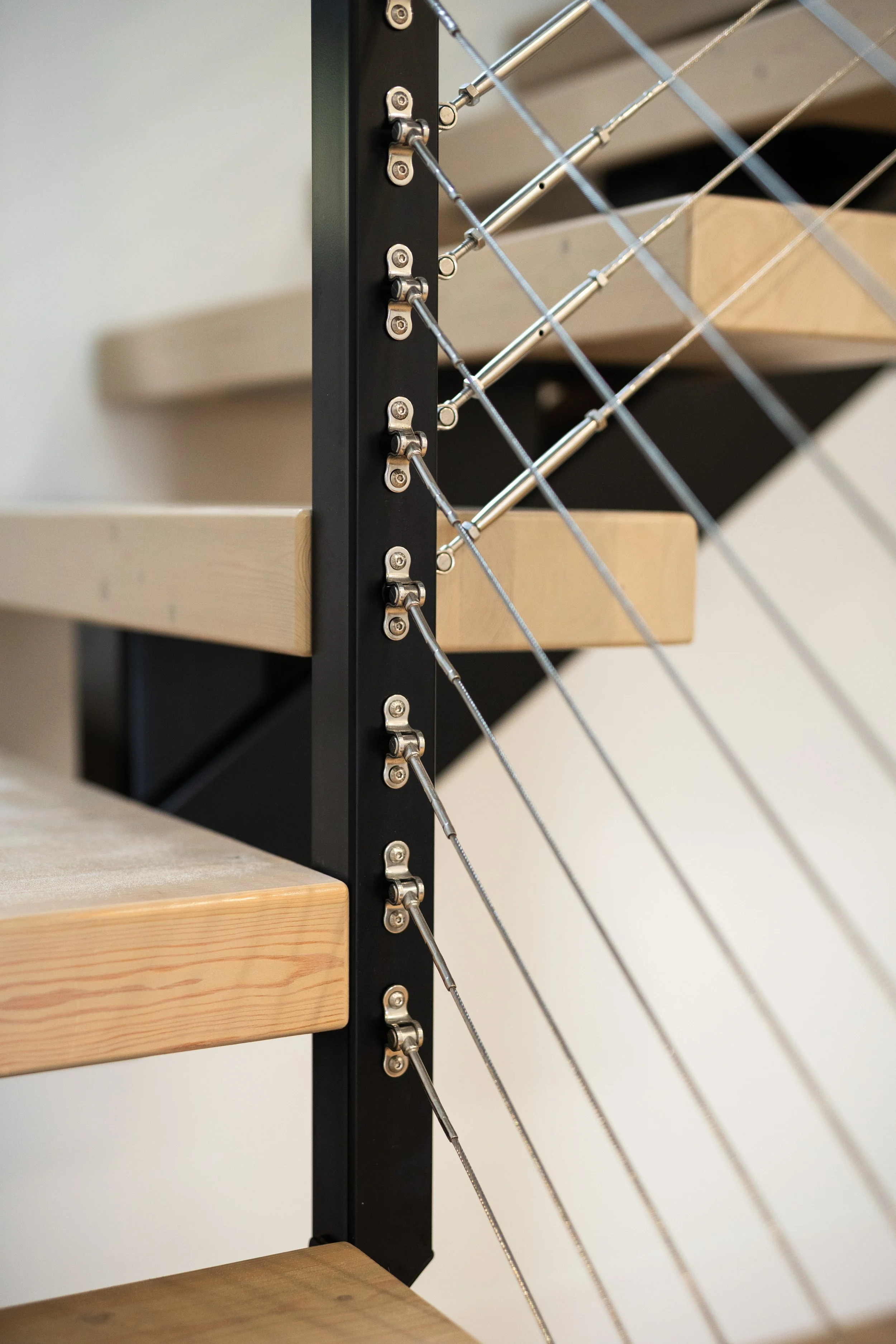 Close-up of a modern stair railing with a black metal support and stainless steel cables, alongside wooden stairs with light-colored wood.