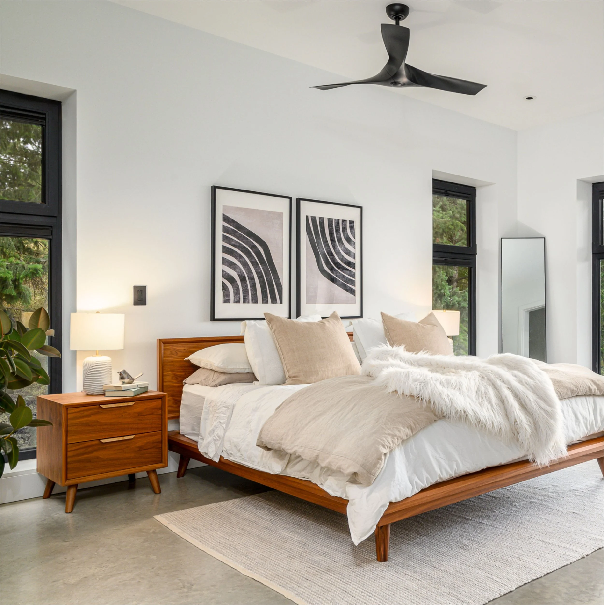 A modern bedroom with a wooden bed frame, white bedding, and beige pillows. There is a matching wooden nightstand with a white lamp and an alarm clock. The room has large black-framed windows, abstract black-and-white artwork on the wall, a gray floo