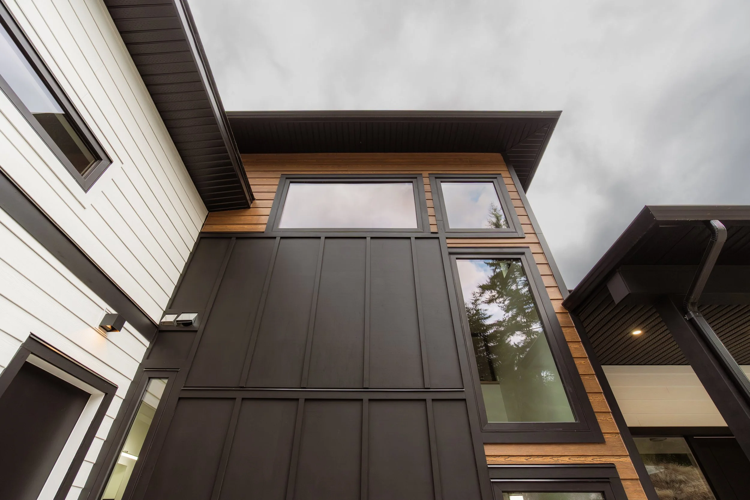 A modern multi-story house with a flat roof, featuring a combination of white, black, and wood-paneled exterior walls, large windows, and a cloudy sky in the background.
