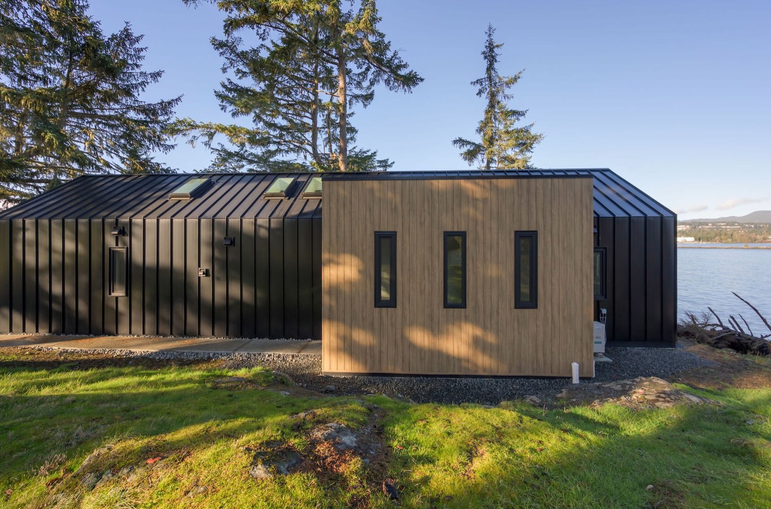 Modern house with black metal exterior and wood accents situated near water, with green grass and tall trees in the background.