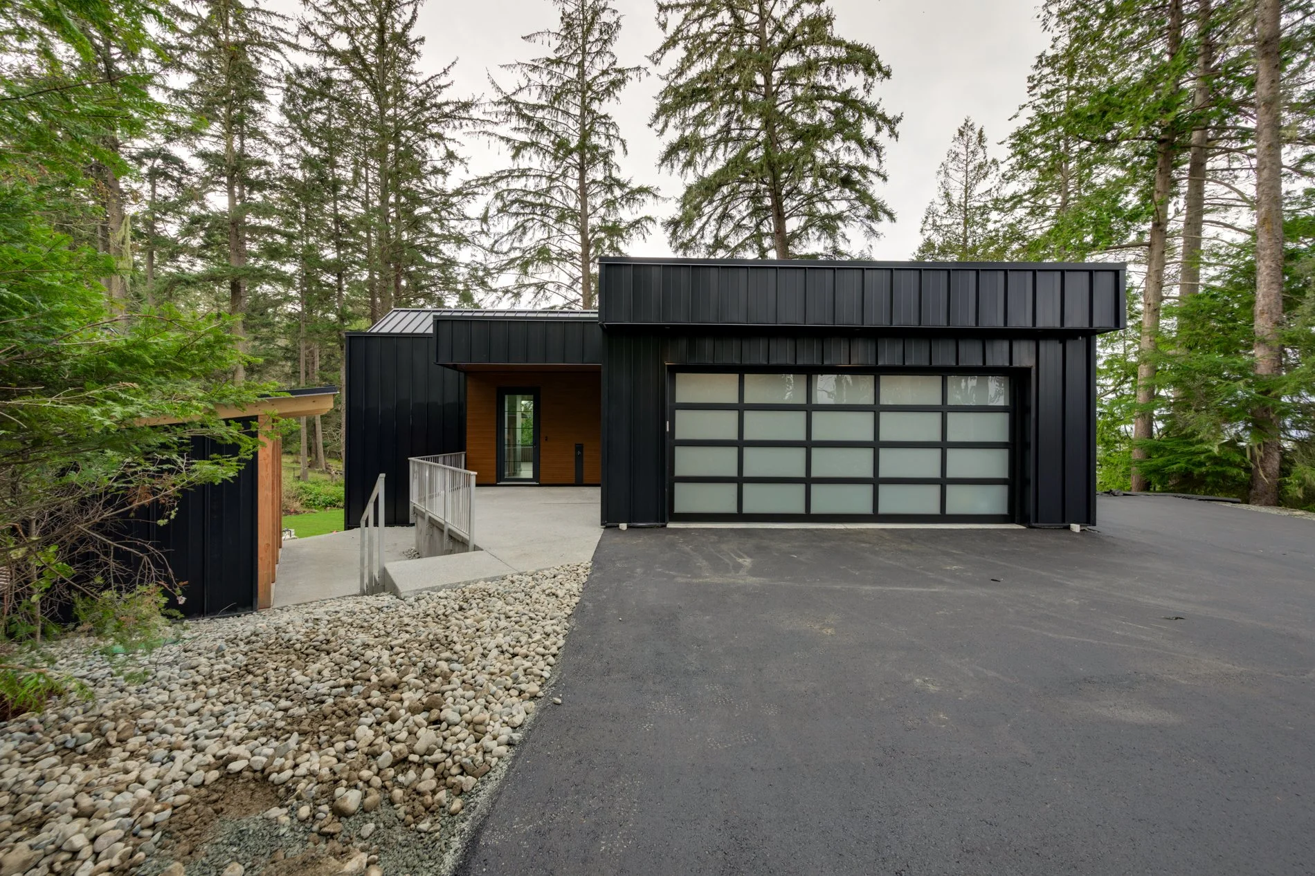 Modern black house with a garage and front entrance, surrounded by trees, concrete pathway, and a gravel area.