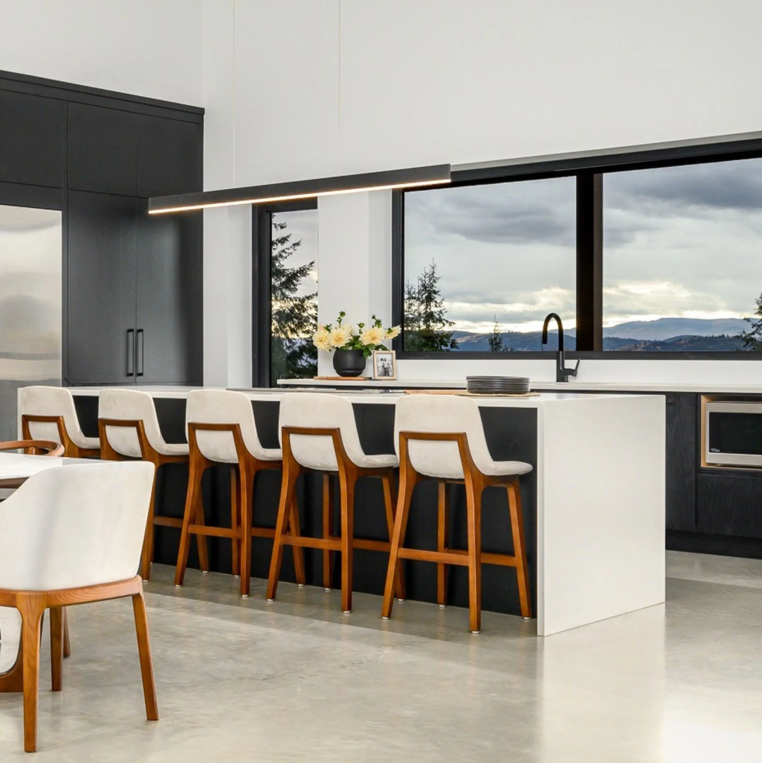 Modern kitchen with white countertops, black cabinets, large window showing cloudy sky and trees, a black vase with white flowers, and a framed photograph on the counter. Dining chairs are visible in the foreground.
