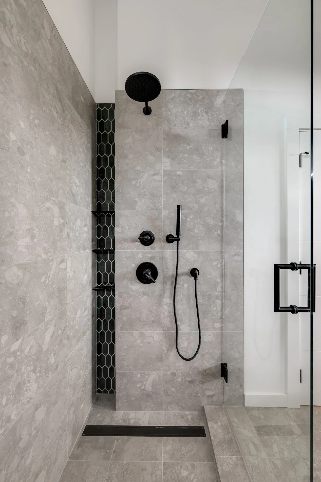 A walk-in shower with grey marble tiles, a black shower head, a handheld shower with a hose, and black fixtures. There is a vertical strip of black hexagonal tiles on the left and a glass door on the right.