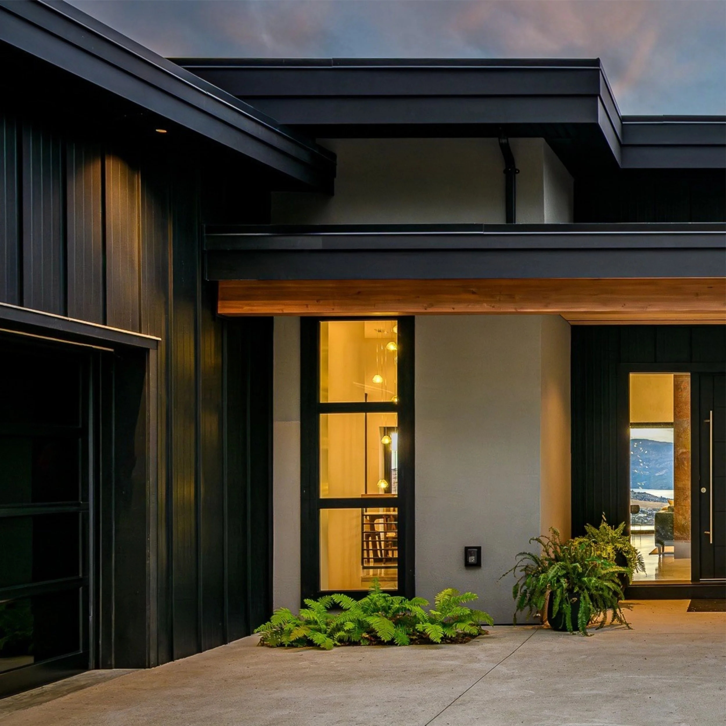 Exterior view of a modern building with black panels and large windows, tropical plants in front, with a sunset sky in the background.