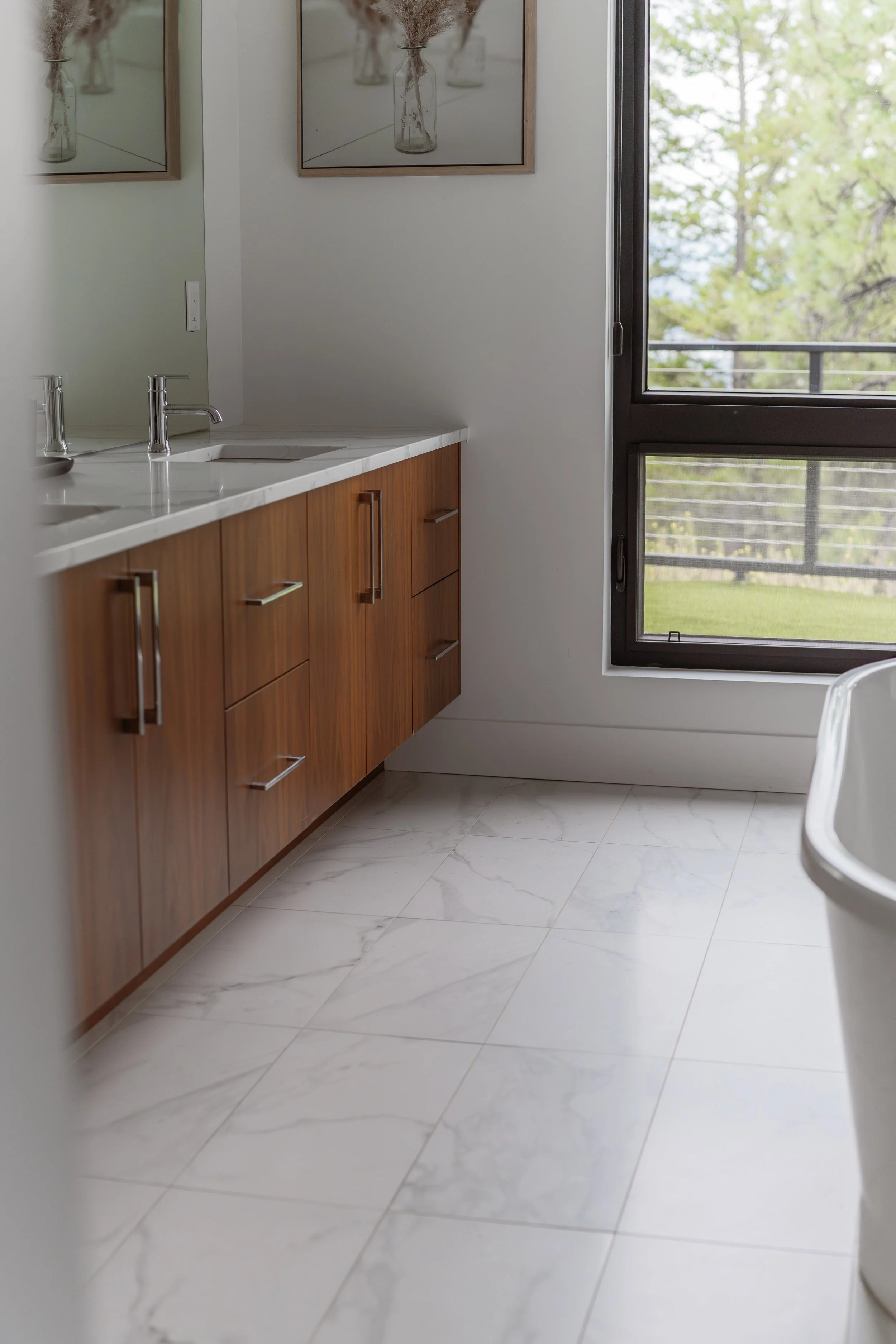 A bathroom with white marble tiled floor, a wooden vanity with a white countertop, twin silver faucets, a mirror, framed artwork with dried flowers, and an open window showing green trees outside.