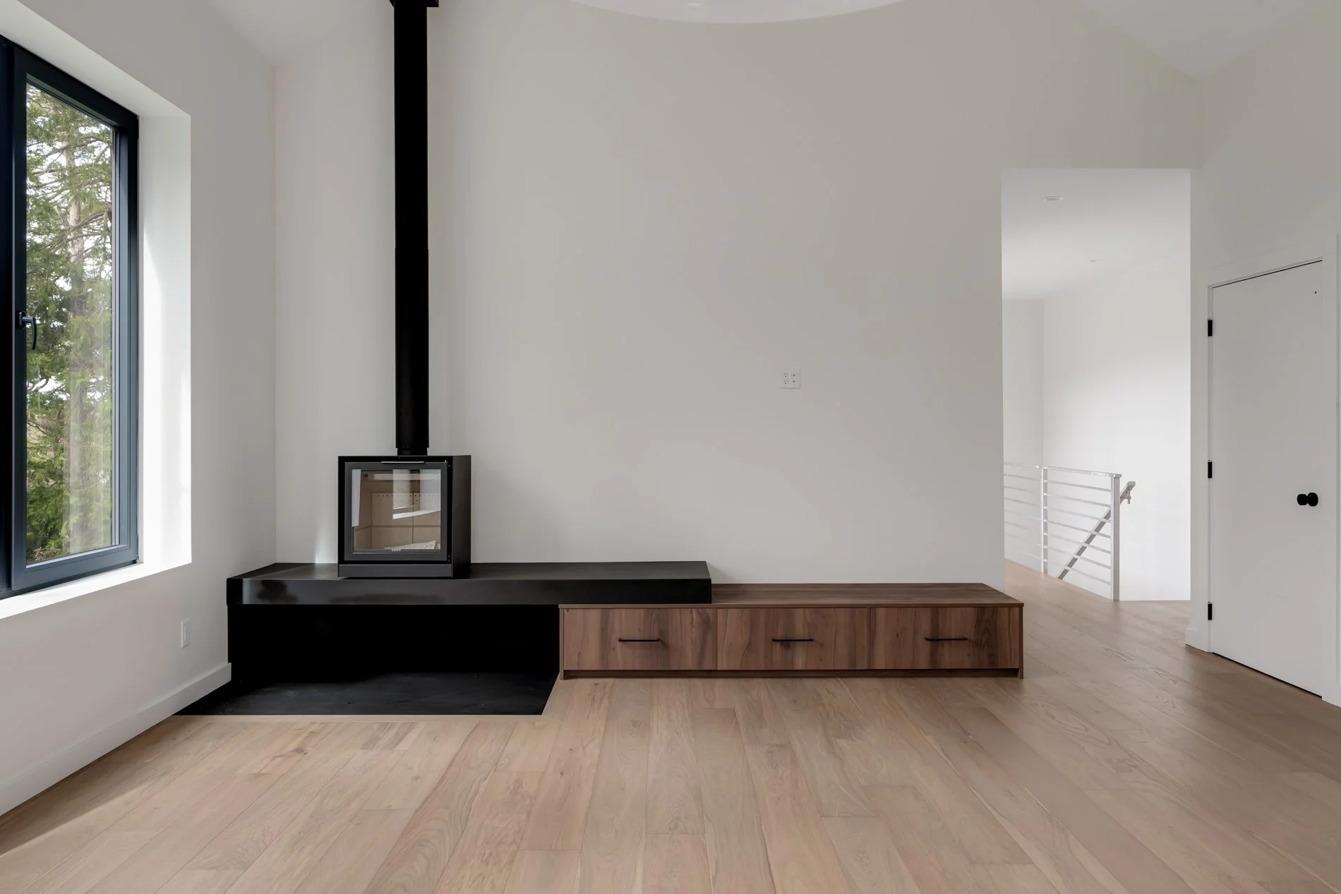Interior of a modern, minimalist living space with a wood stove on a black platform, a large window showing trees outside, light-colored hardwood floors, and white walls. Part of a staircase and a white door with a black handle are visible.