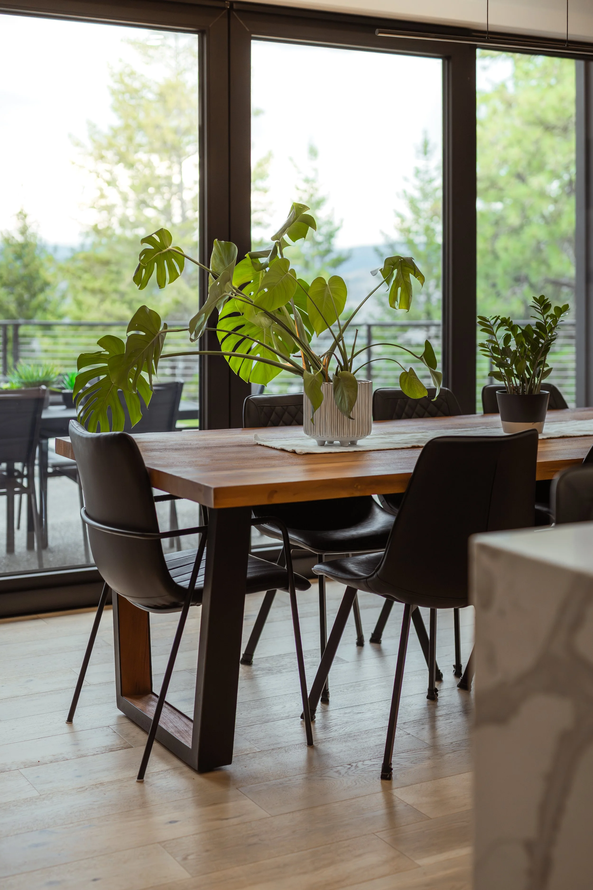 Interior view of a dining area with a wooden table, black chairs, and potted plants, large windows with a view of green trees outside, and a light-colored wooden floor.