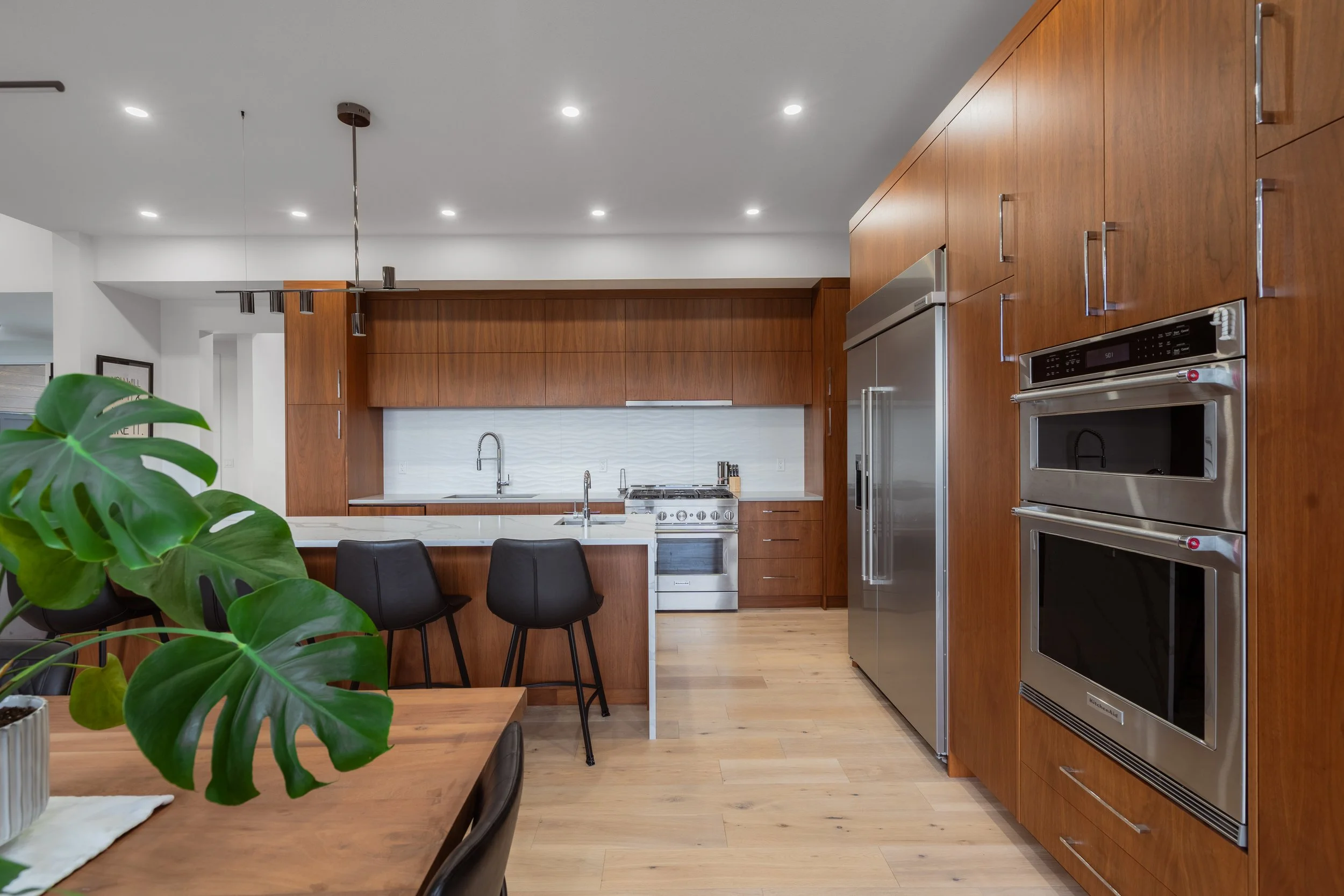 Modern kitchen with wooden cabinets, stainless steel appliances, and a dining table with black chairs. Green plant in the foreground, pendant lights, and recessed ceiling lights.