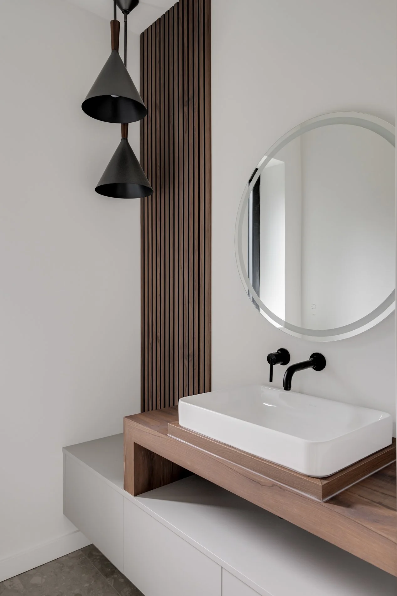 Modern bathroom with a rectangular white vessel sink on a wooden vanity, black fixtures, a round mirror, vertical wood paneling, black pendant lights, and a white storage cabinet below.