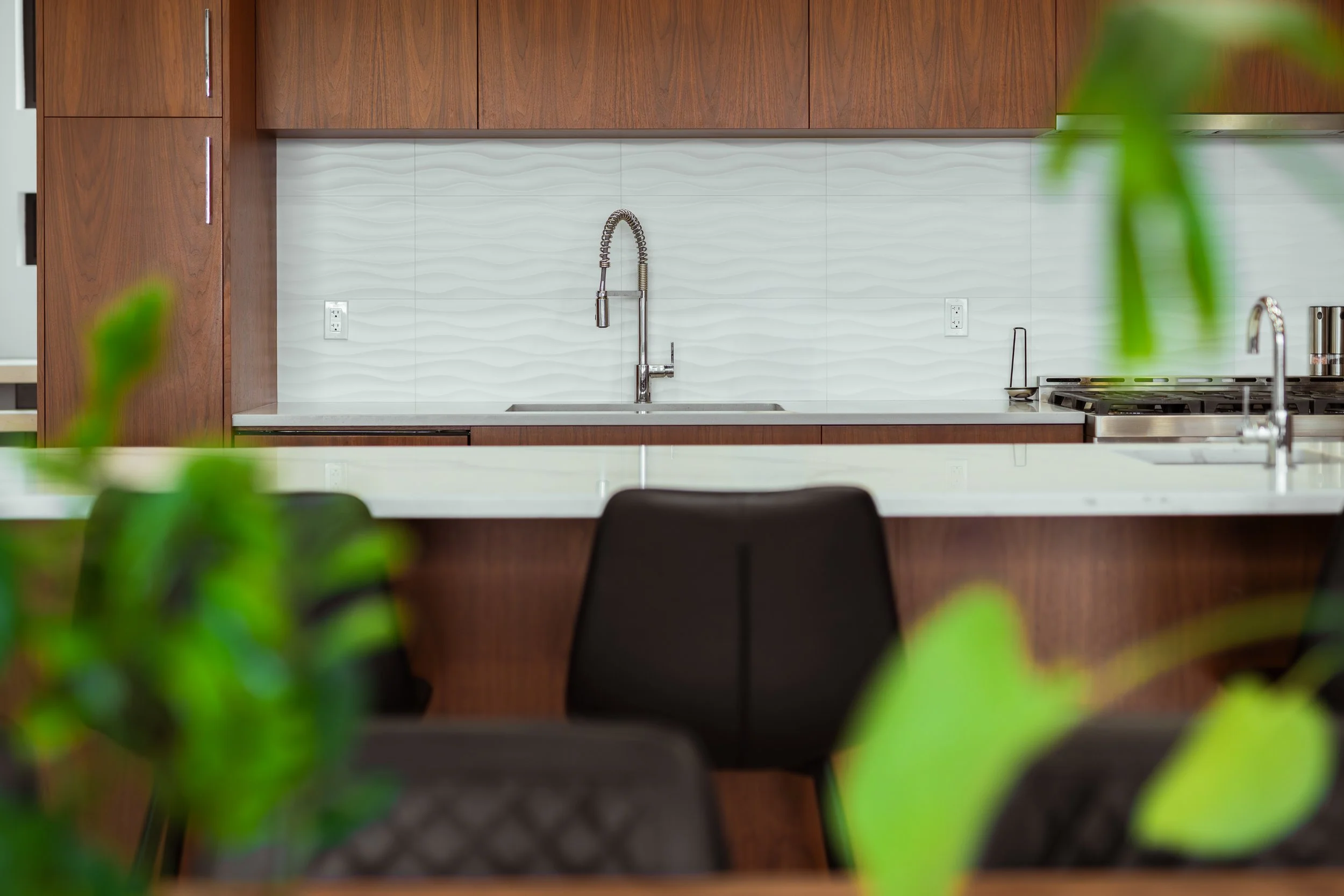 Modern kitchen with white textured backsplash, dark wood cabinets, stainless steel faucet, and black chairs in the foreground.