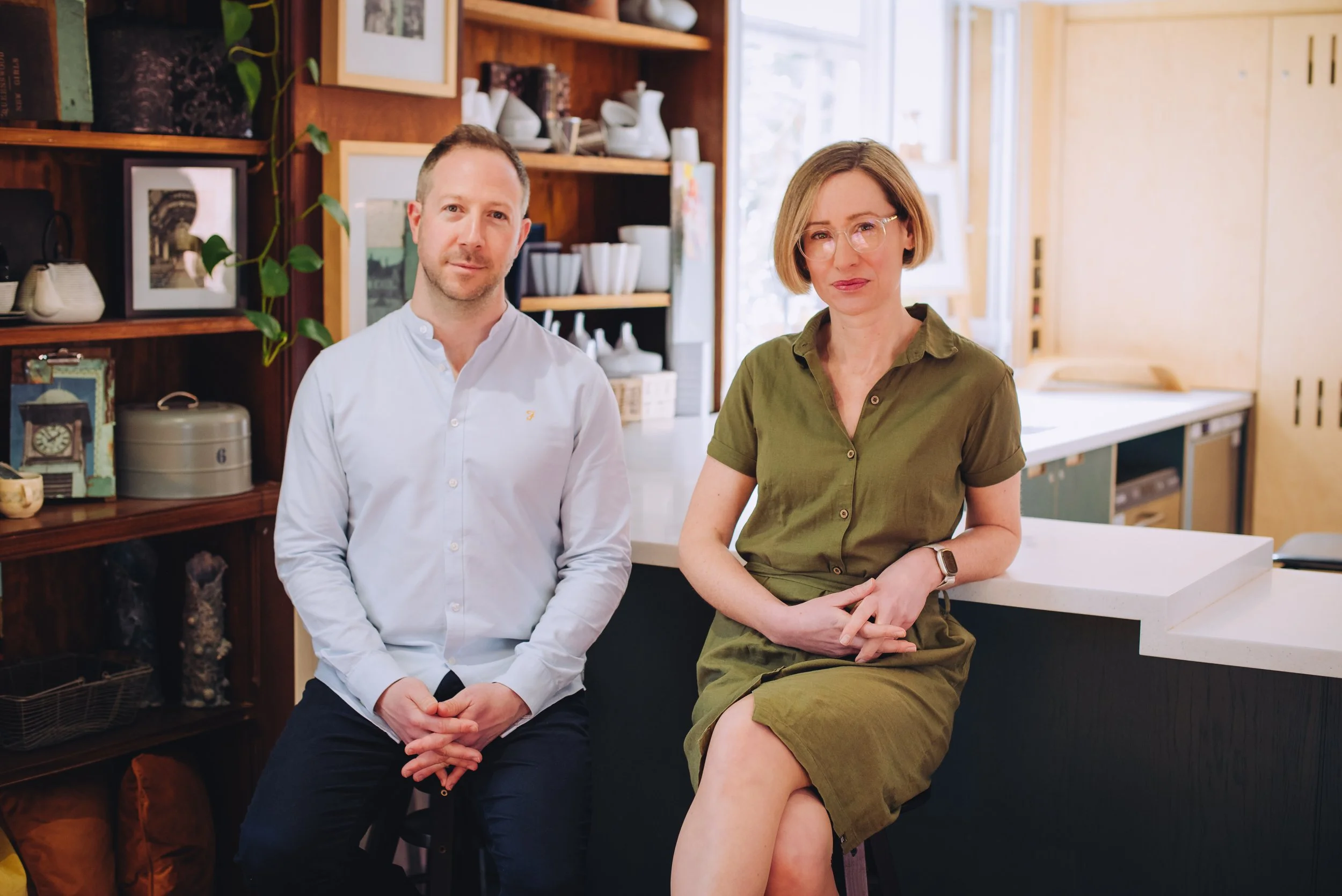 A man and a woman sitting in a cozy, brightly lit room with wooden shelves and a large window behind them. The man is wearing a white shirt and the woman is wearing an olive green dress with glasses.
