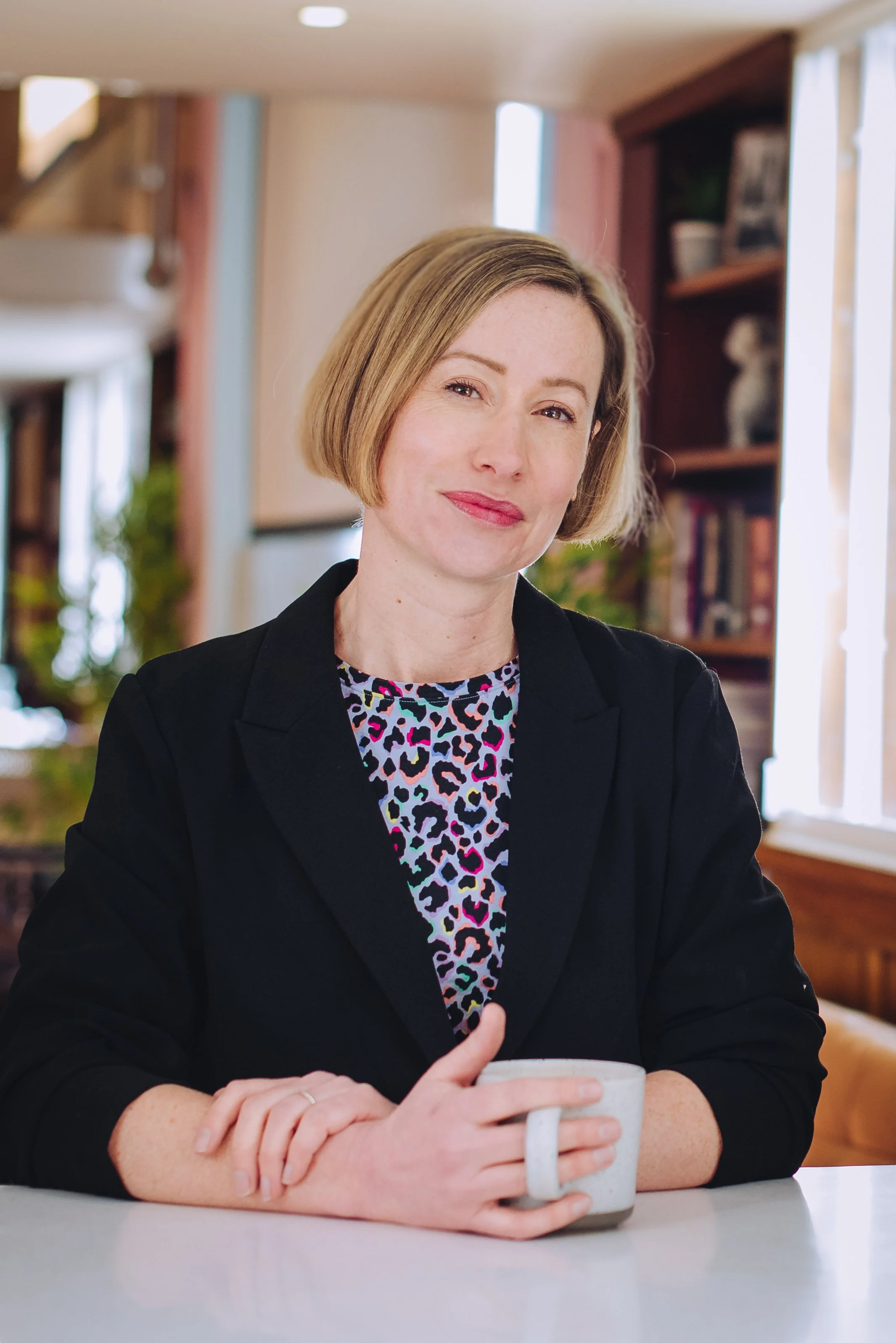 A woman with short blonde hair, wearing a black blazer over a colorful animal print top, sitting at a table with a white mug in her hands, in a cozy indoor space with bookshelves and natural light.