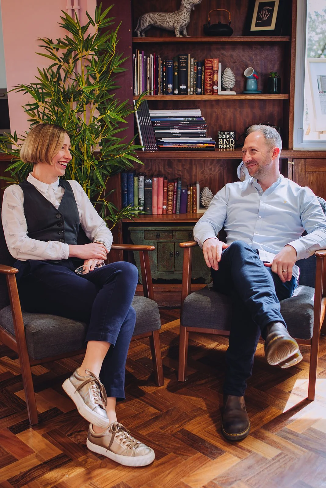 A man and a woman are sitting in chairs, smiling and having a conversation in a cozy room. There is a bookshelf behind them with various books and decorative items, including a framed quote that says "Breathe Deeply and Repeat." The room has wooden flooring, a large green plant, and a window with natural light.