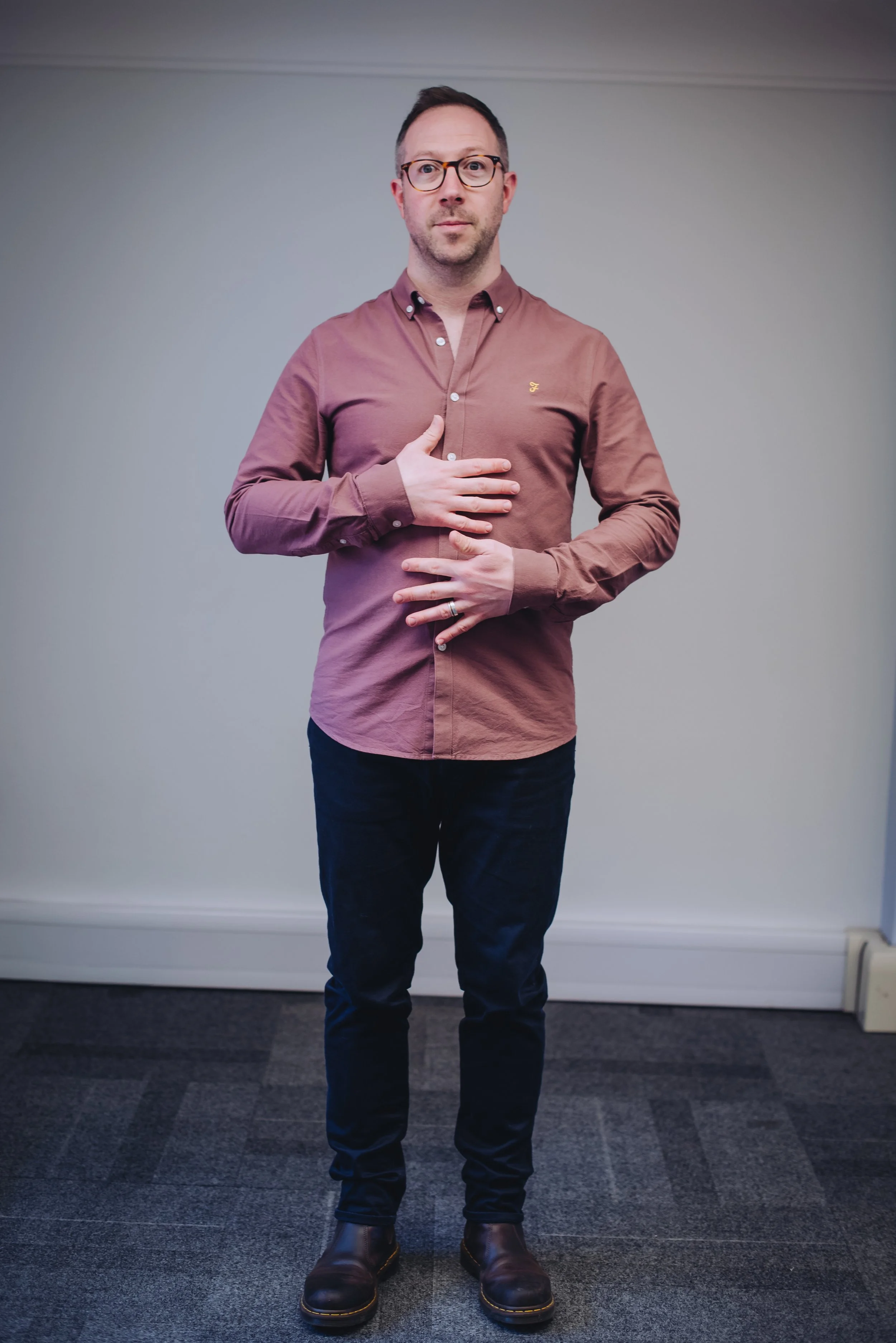 A man wearing glasses, a button-up shirt, dark jeans, and leather boots stands in front of a plain wall. He is holding his right hand over his chest and his left hand over his stomach.