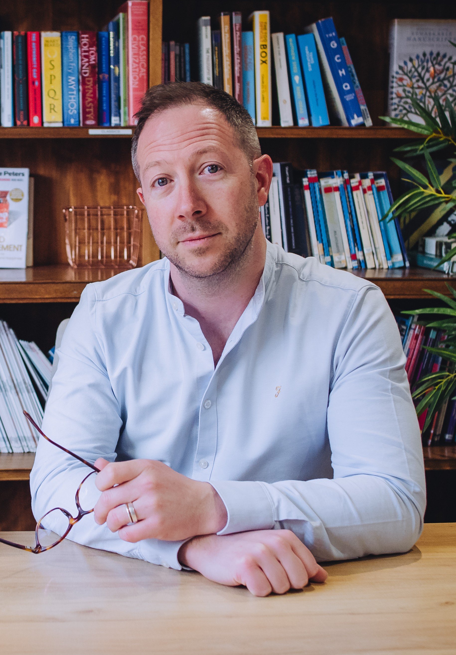 A man with short brown hair and light skin sitting at a wooden table, holding glasses with both hands, against a background of a bookshelf filled with colorful books and a potted plant.