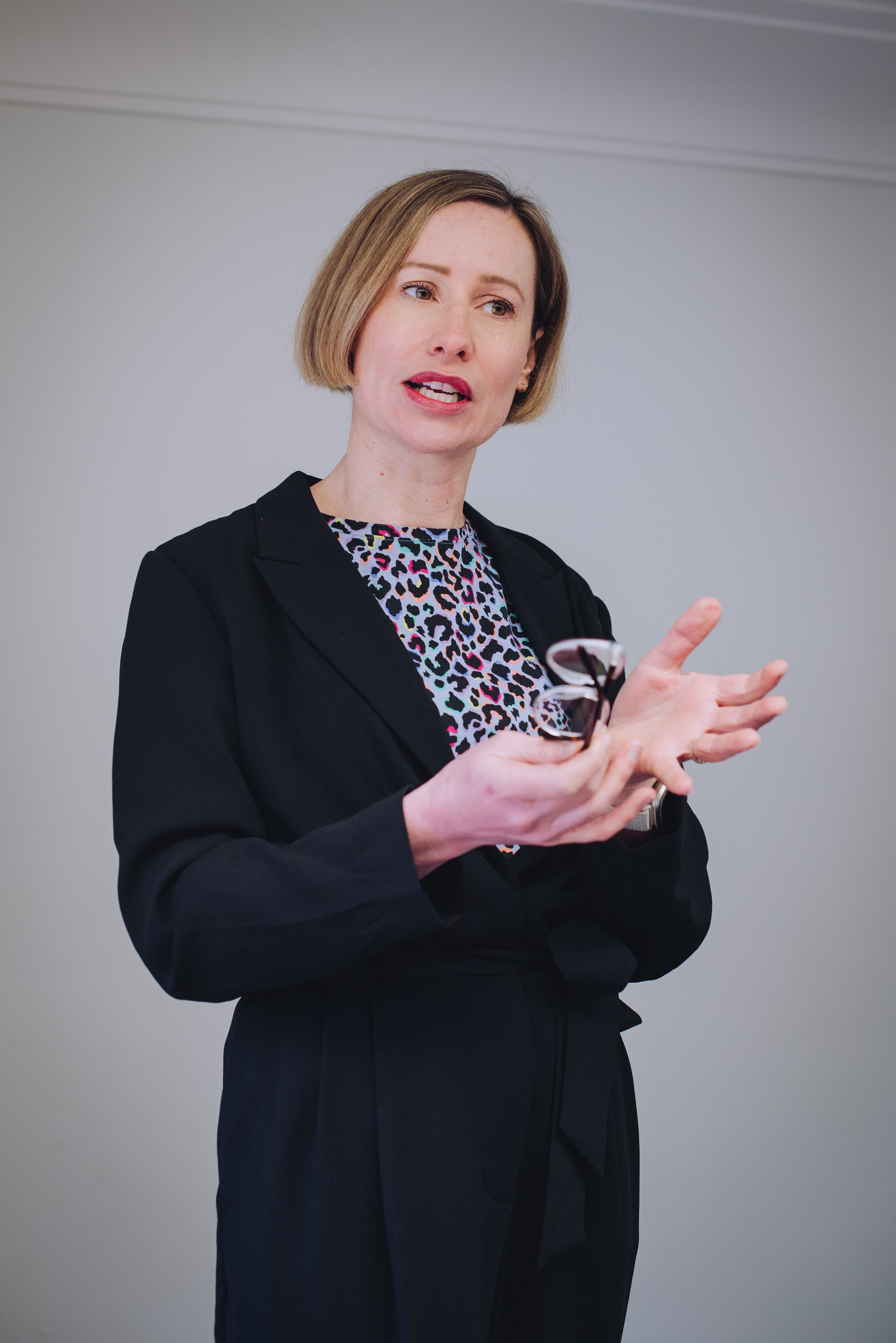 A woman with a short, blonde bob haircut and fair skin is speaking and gesturing with her hands. She is wearing a black blazer over a patterned blouse with animal prints in bright colors. She is standing against a plain, light-colored wall.