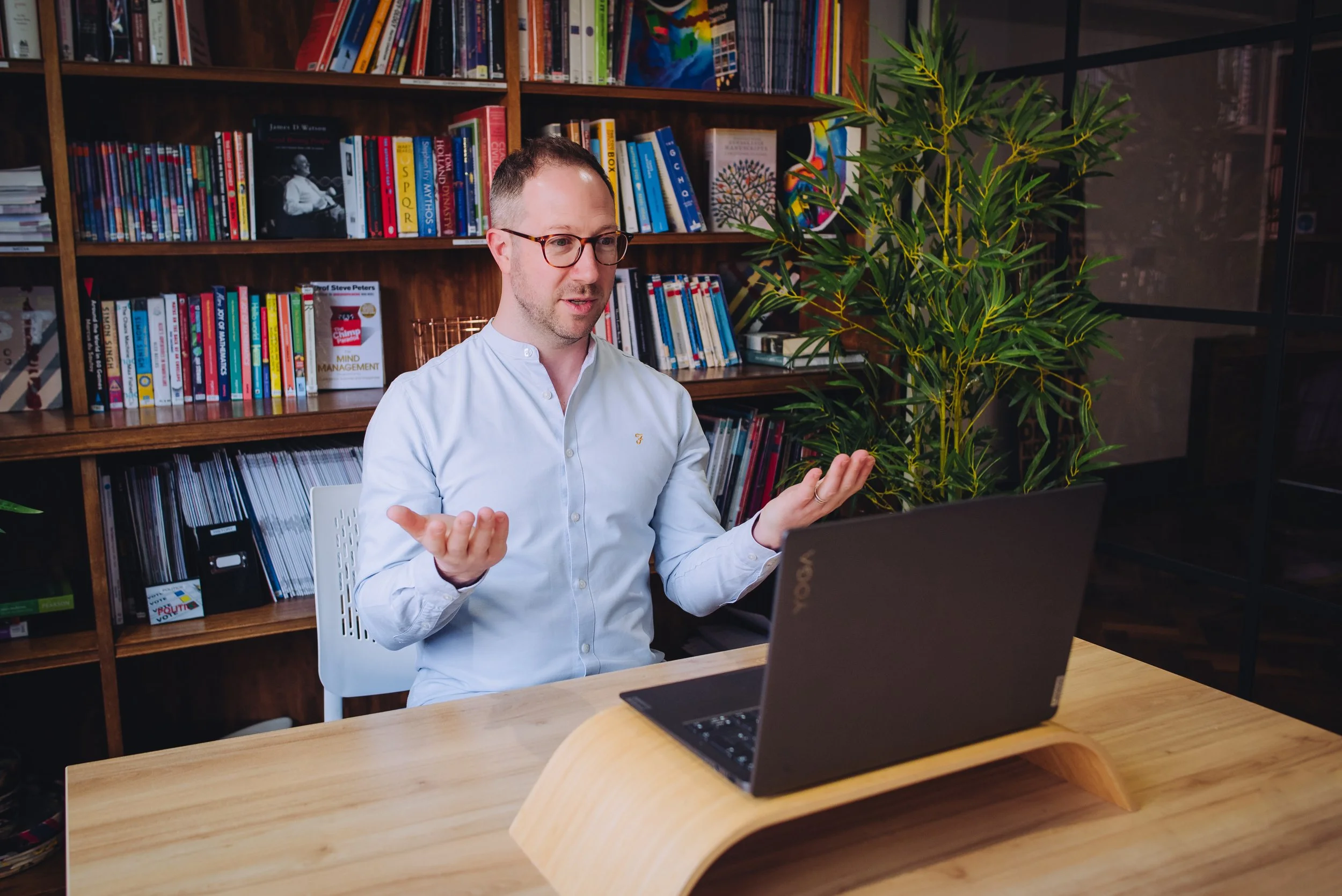 A man with glasses and a white shirt sitting at a desk with a laptop, gesturing with his hands, in a room with a wooden bookshelf filled with books and colorful artwork, and a large green plant nearby.
