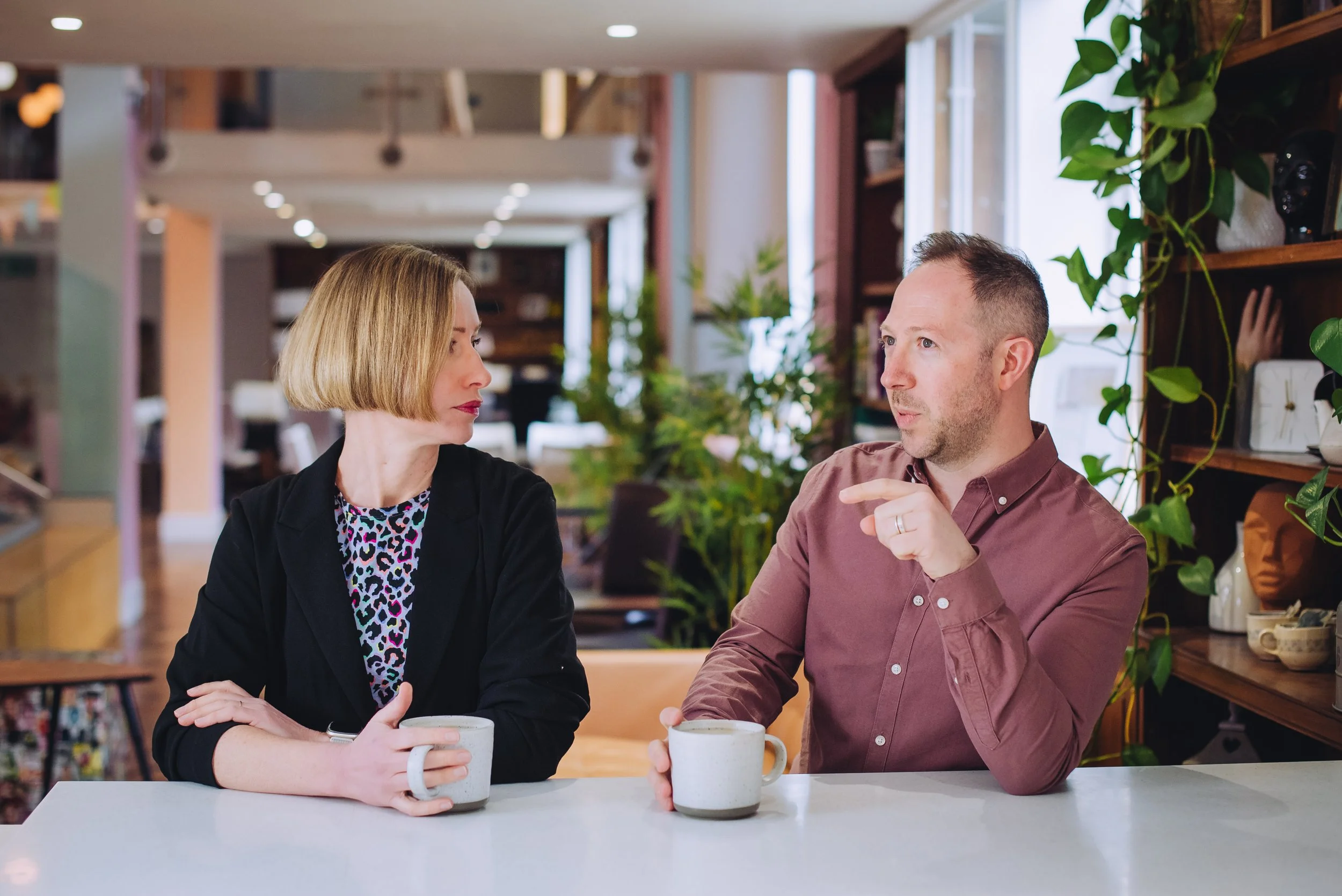 A man and a woman sitting at a table in a cozy cafe, each holding a white mug, engaged in conversation with indoor plants and wooden shelves in the background.