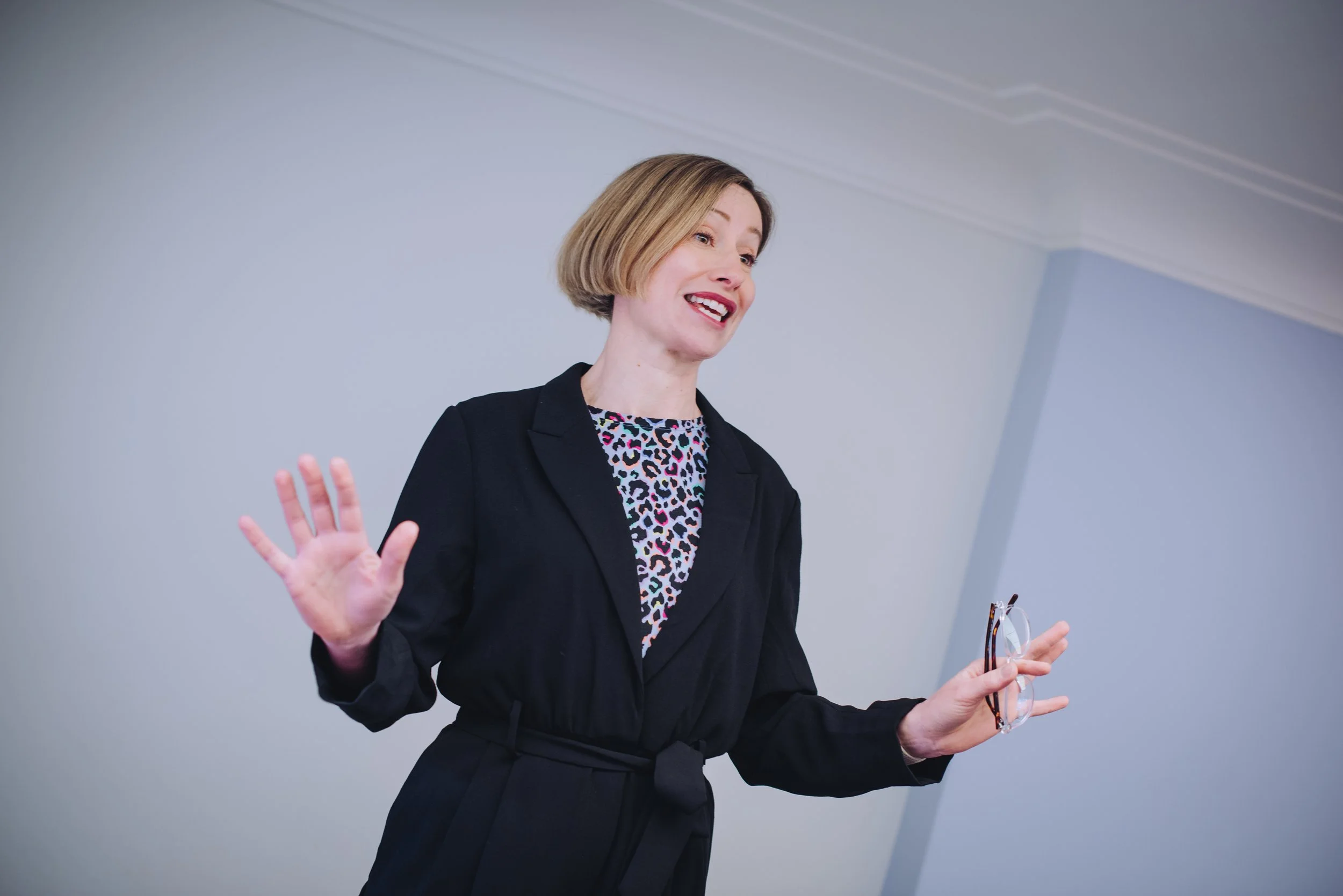 A woman with a short blond bob haircut, wearing a black blazer and leopard print top, is speaking or presenting, with one hand raised and the other holding glasses, against a light gray background.