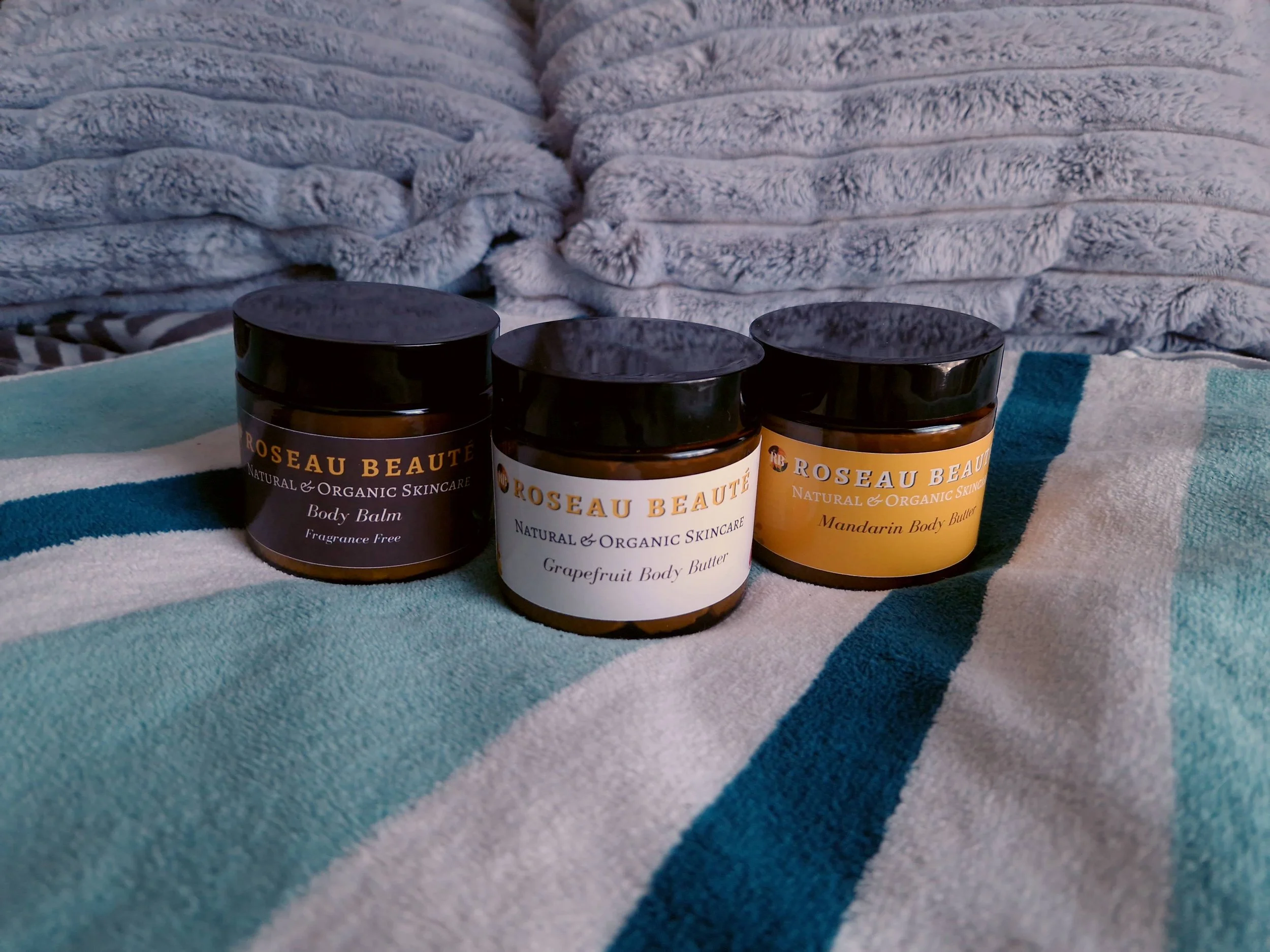 Three jars of Roseau Beauté natural organic skincare products on a striped blue and white towel, with fluffy grey pillows in the background.