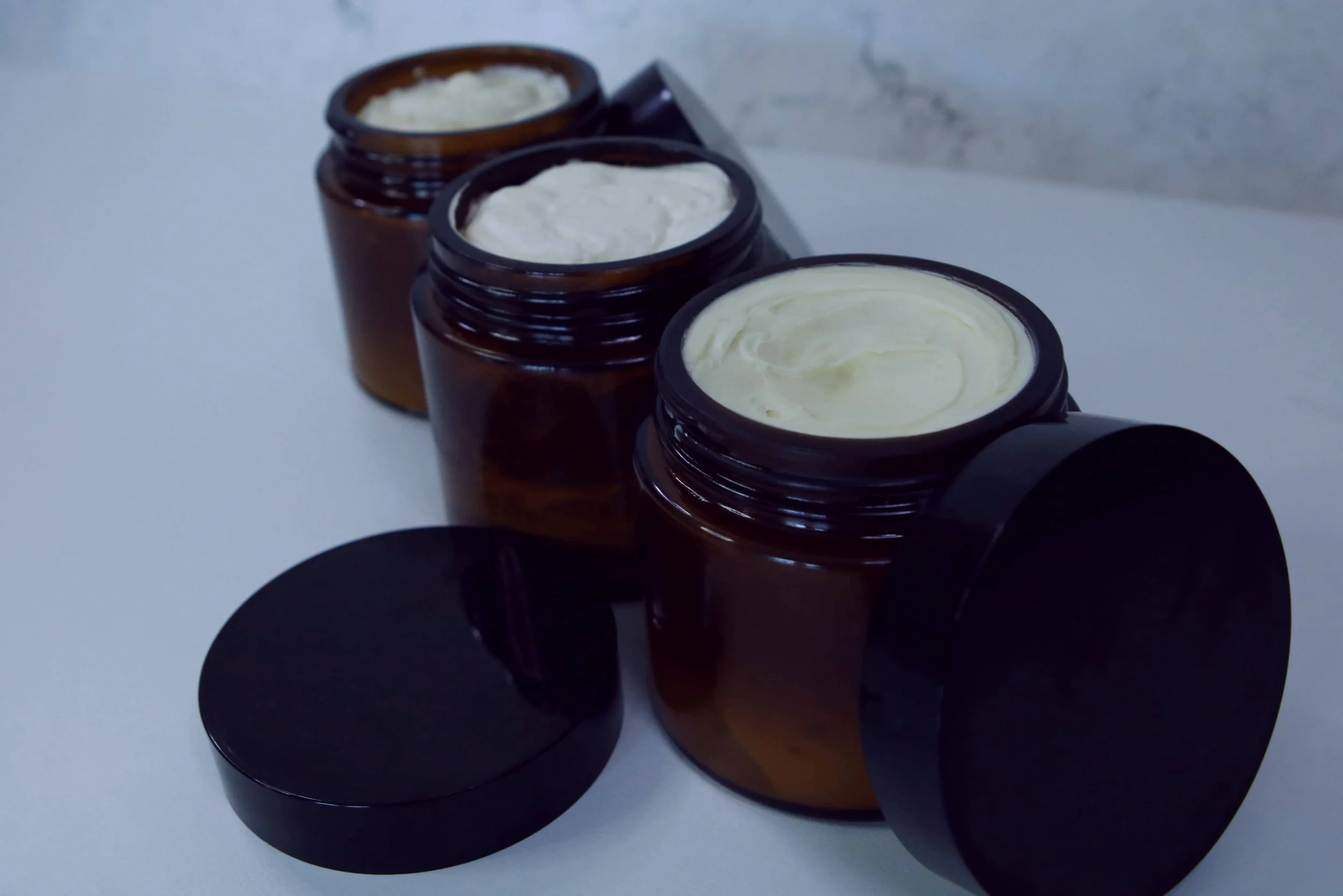 Three open brown glass jars with body butter inside, arranged in a diagonal line on a white surface, with black circular lids placed in front of or near each jar.
