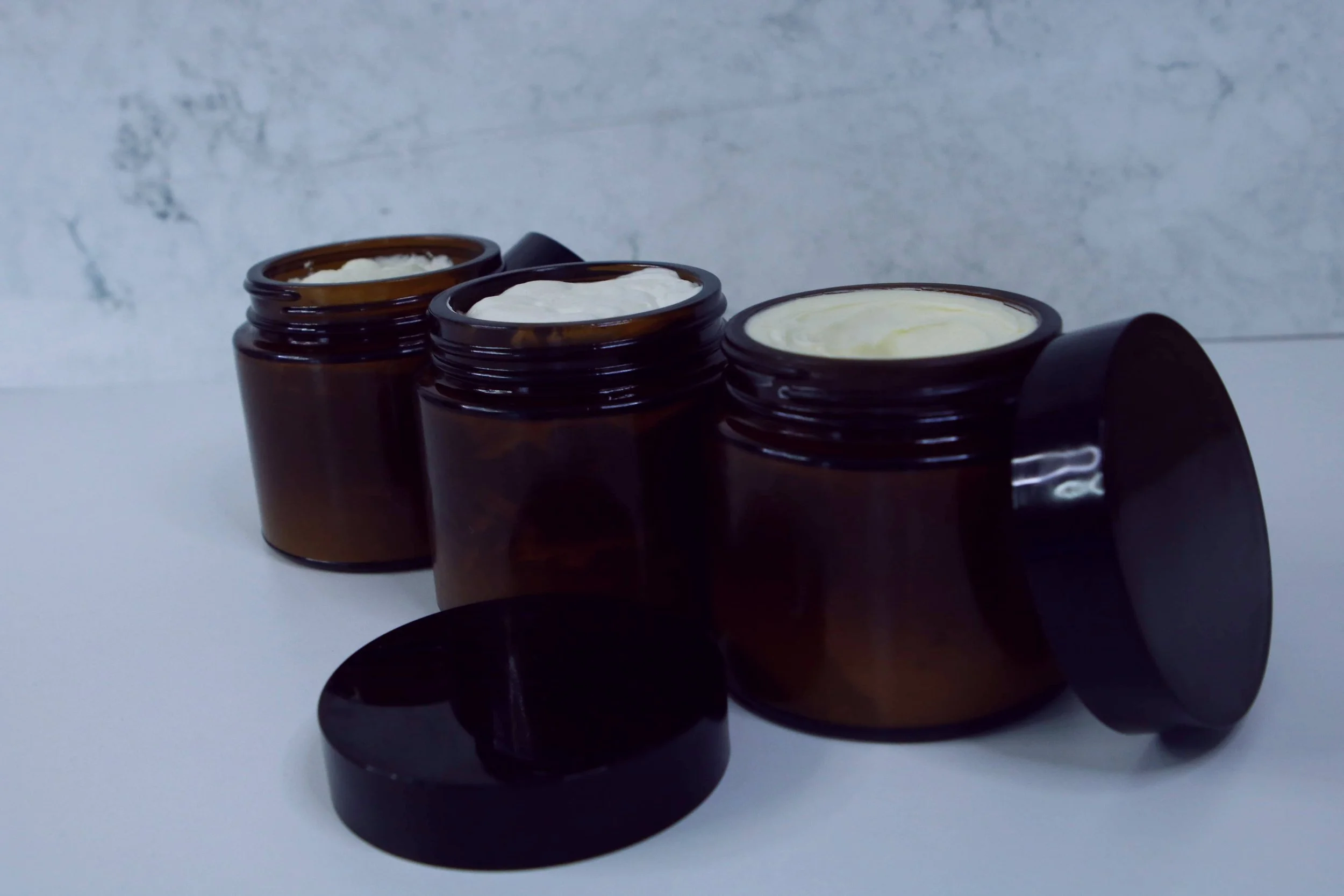 Three open brown glass jars with body butter, placed on a white surface against a light grey background.
