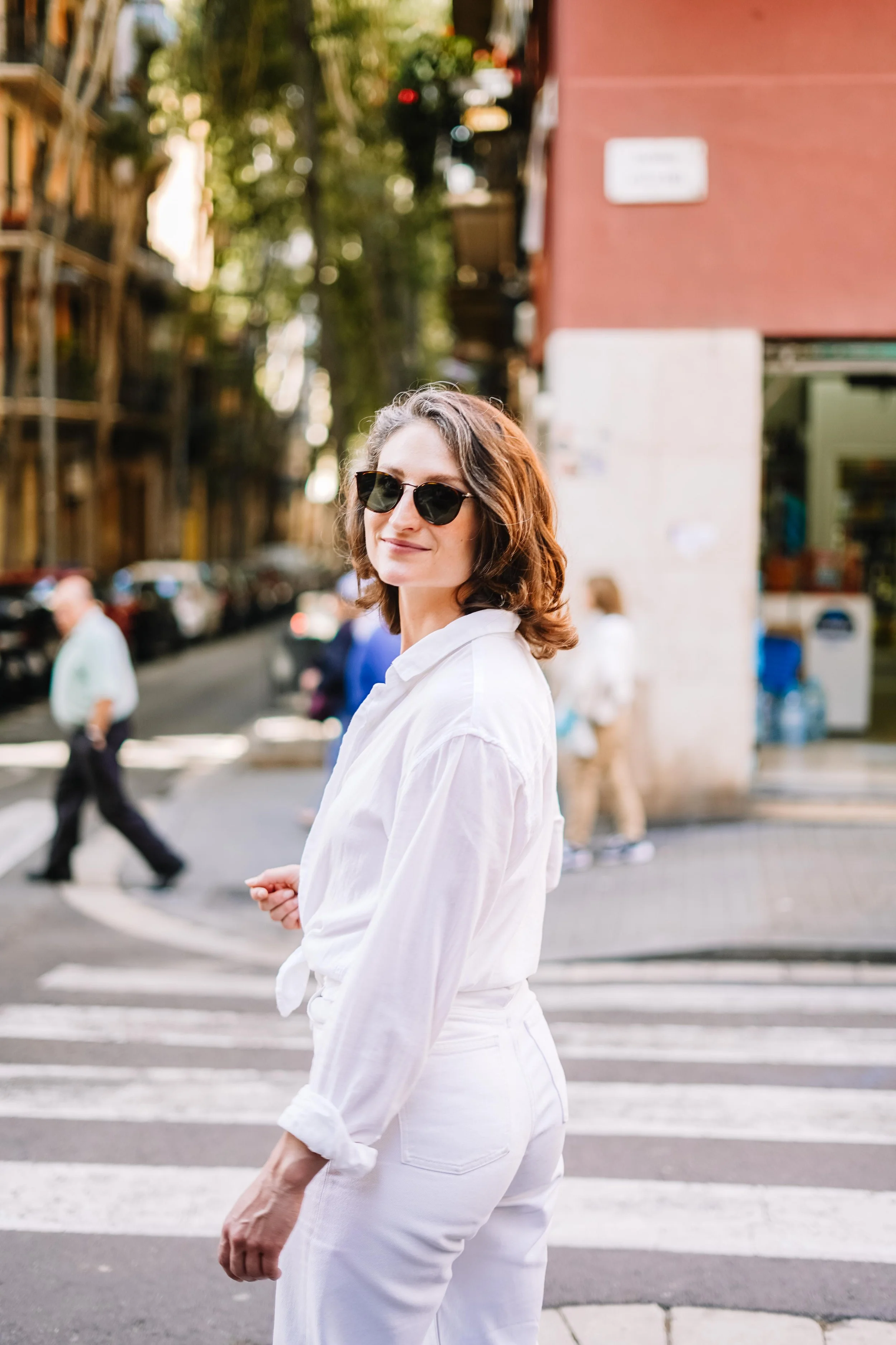 A woman (Casey Berglund) with wavy brown hair, wearing dark sunglasses, a white shirt, and white pants, standing on a city street at a crosswalk with blurred pedestrians and cars in the background.