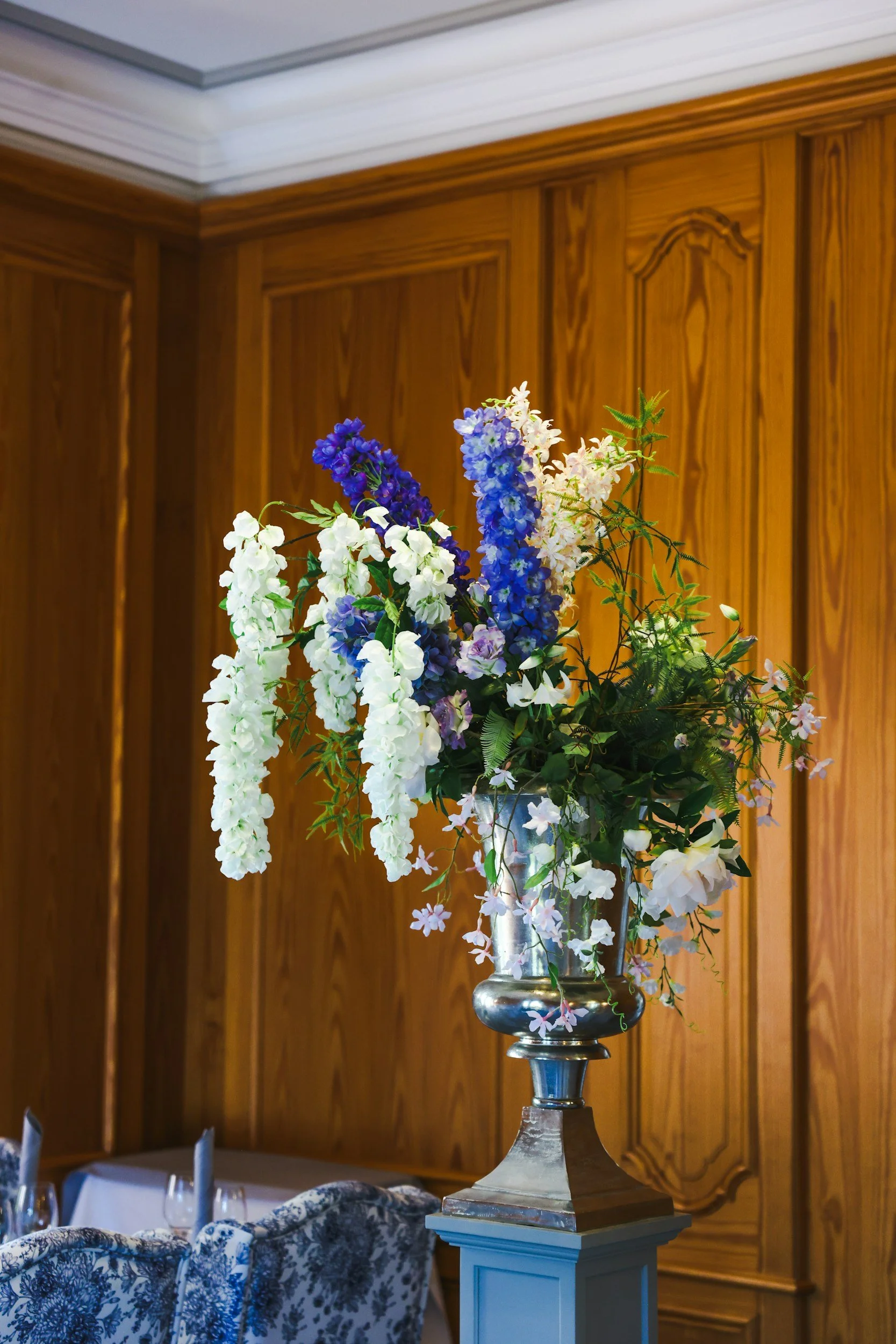A bouquet of white, purple, and blue flowers in a tall silver vase on a white pedestal, in front of a wooden paneled wall.