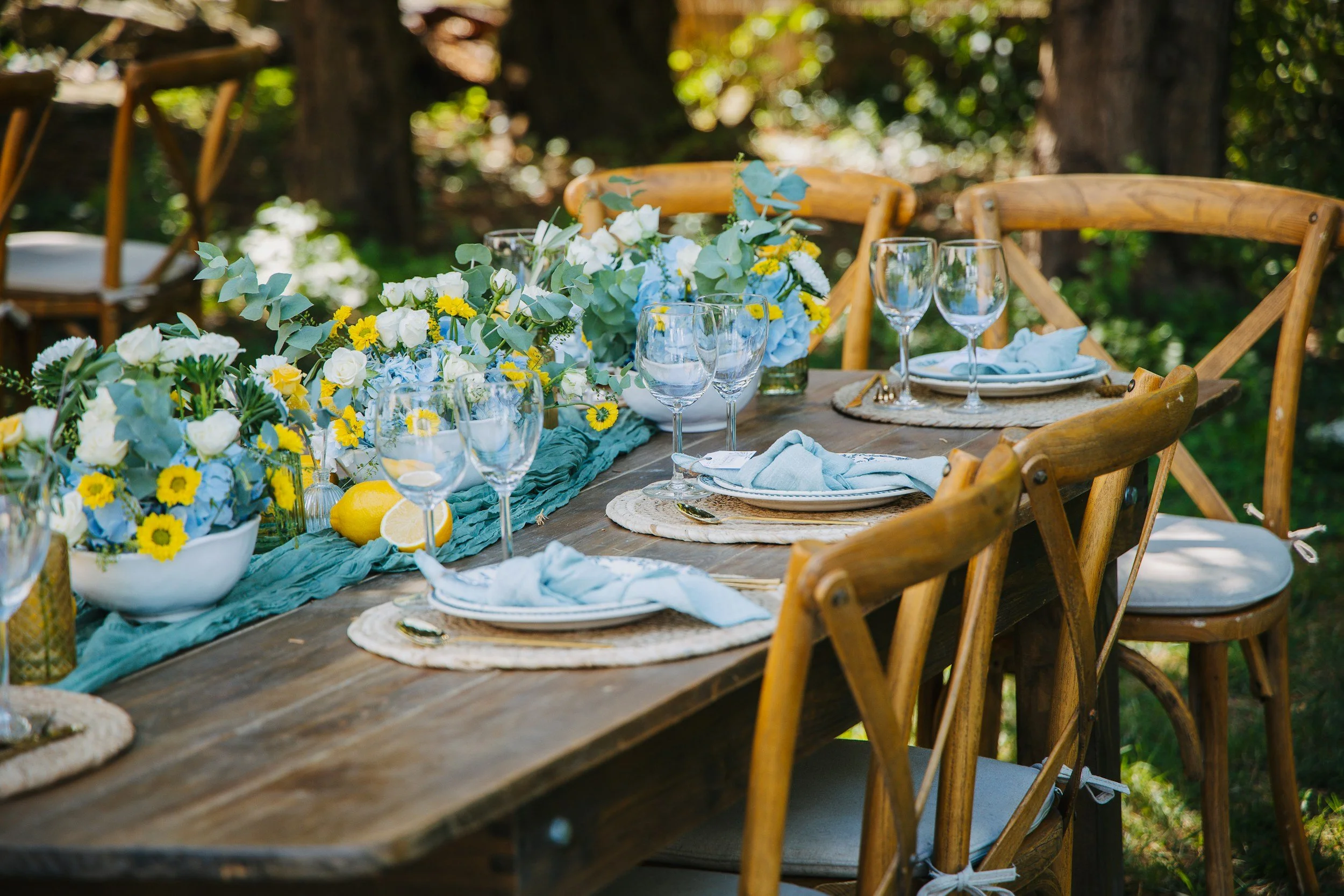 A rustic outdoor table decorated with a floral centerpiece of white, yellow, and blue flowers, set with wine glasses, plates, and napkins. Surrounding the table are wooden chairs and lush green trees in the background, suggesting a garden or backyard setting for a celebration or gathering.