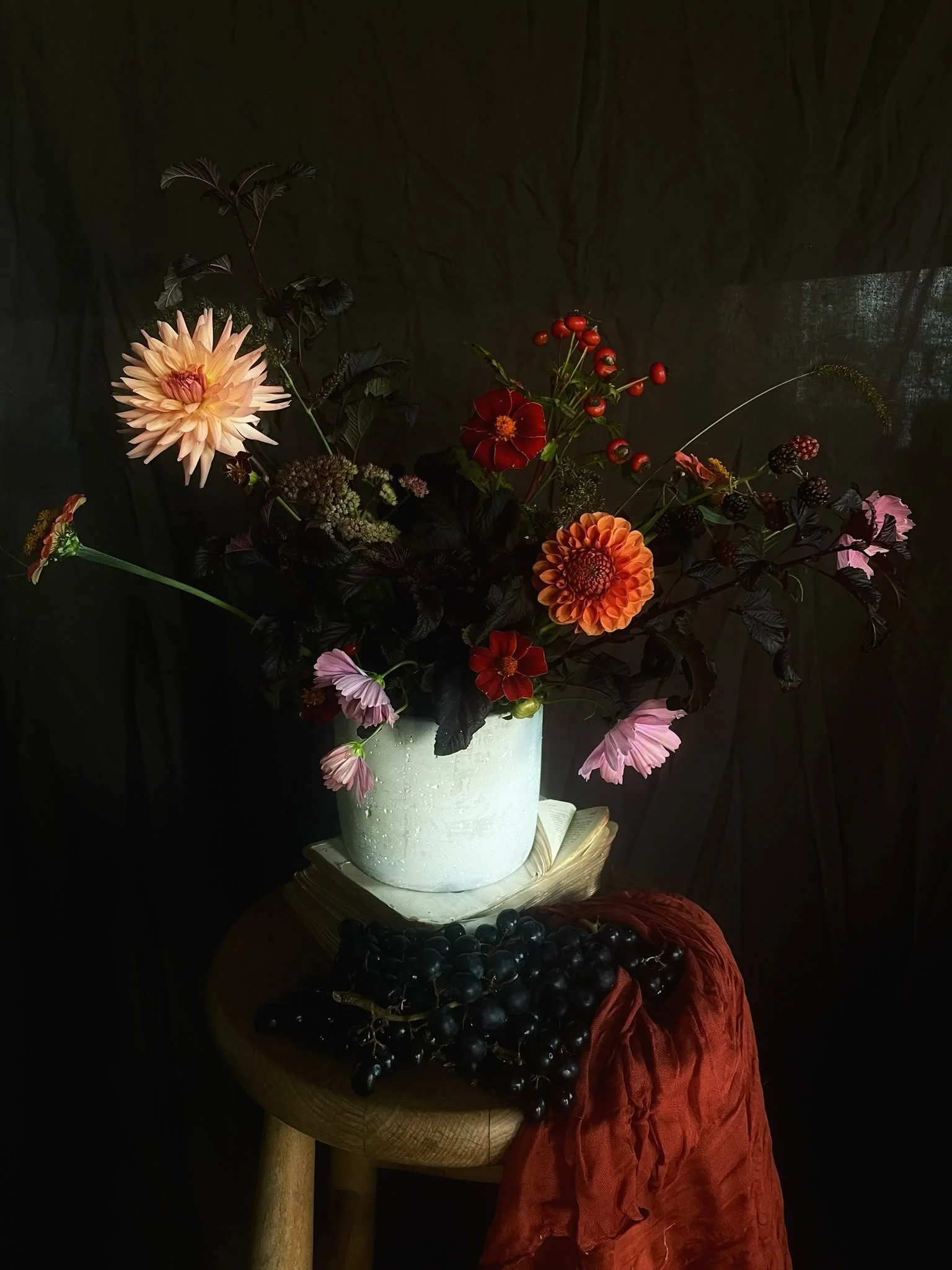 A bouquet of colorful flowers in a white vase on a wooden table, with a bunch of black grapes and a red cloth nearby, against a dark background.
