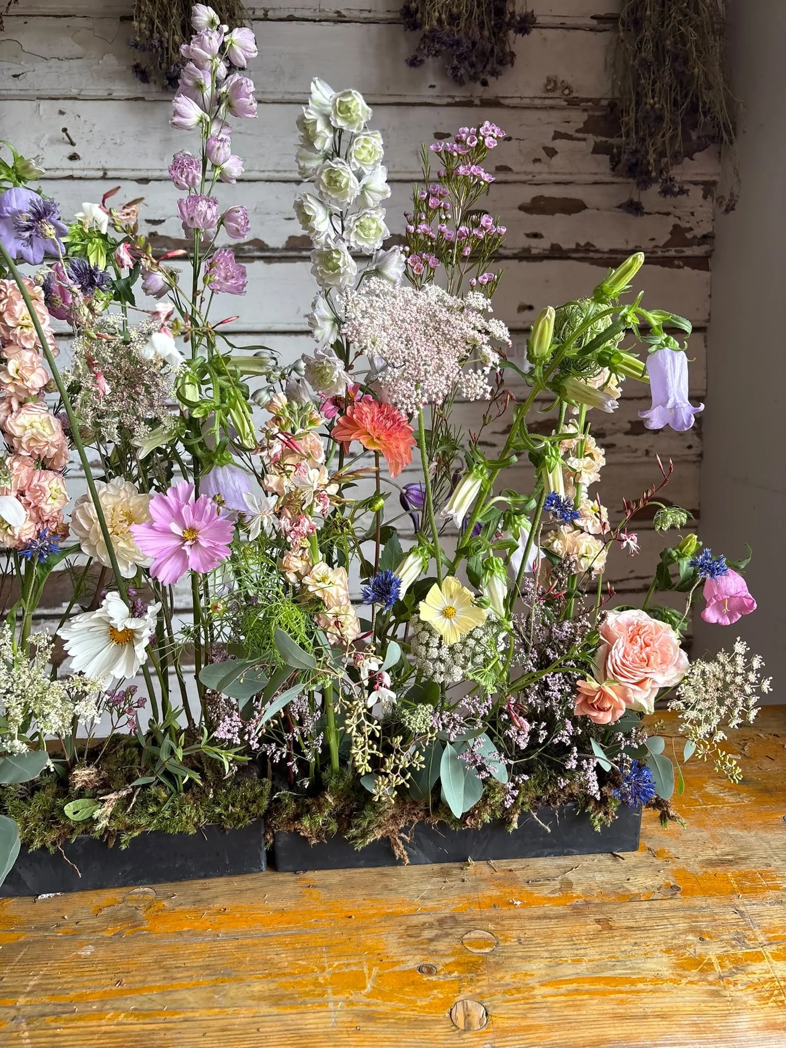 A large mixed flower arrangement in a black rectangular container on a rustic wooden table with a weathered white wooden wall in the background. The arrangement includes pink, white, purple, and cream flowers, with green foliage and some dried flowers hanging on the wall.