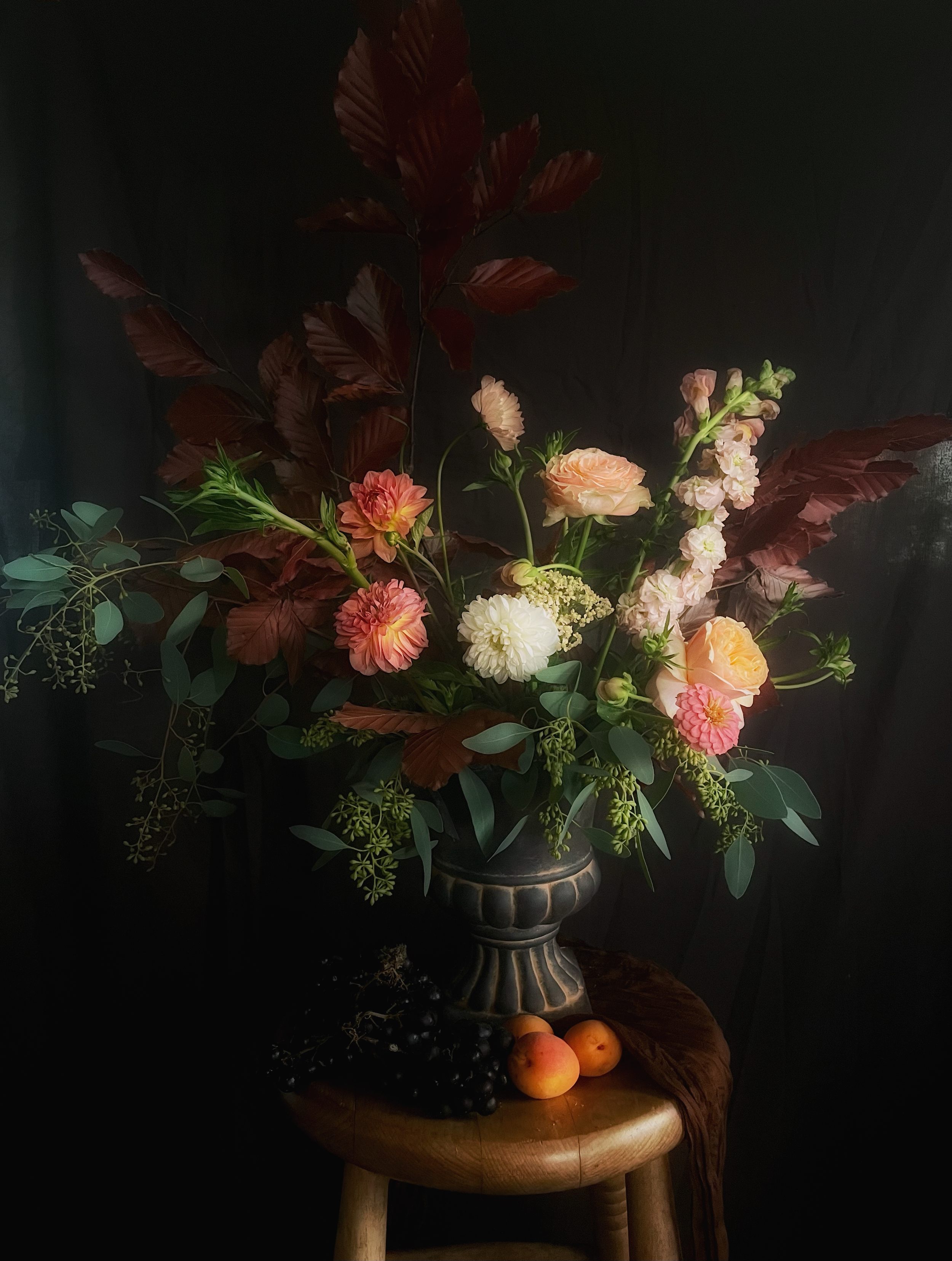 A still life floral arrangement in a decorative vase on a wooden stool with peaches and black grapes at the base, set against a dark background.