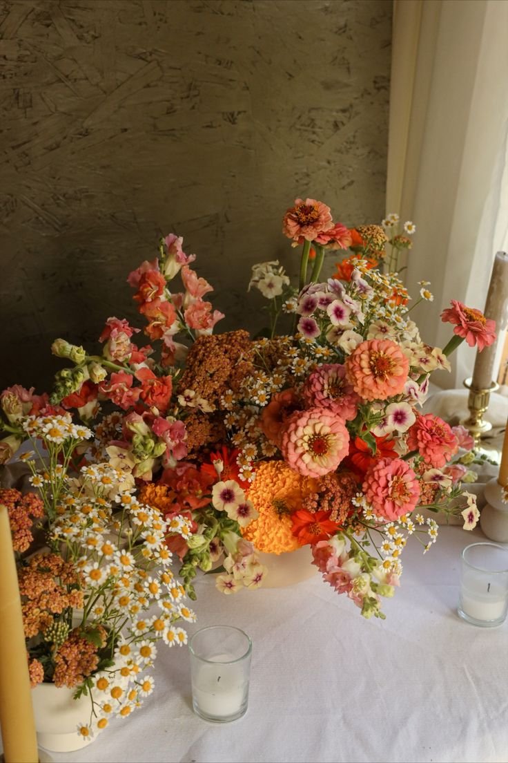 A floral arrangement with pink, orange, and white flowers on a white table with candles.