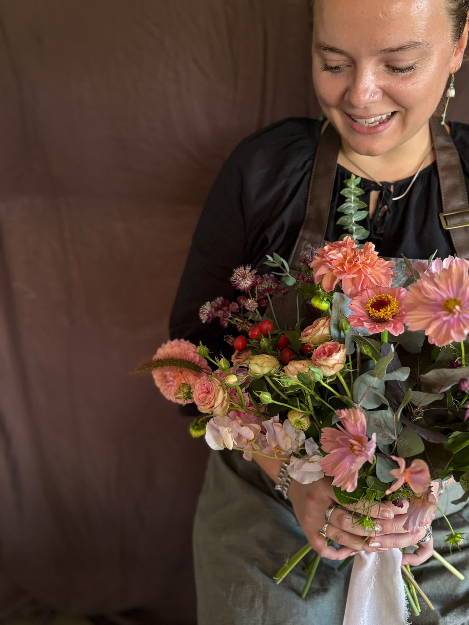 Person holding a colorful bouquet of flowers, including pink, peach, and white blooms, with green foliage, against a brown background.