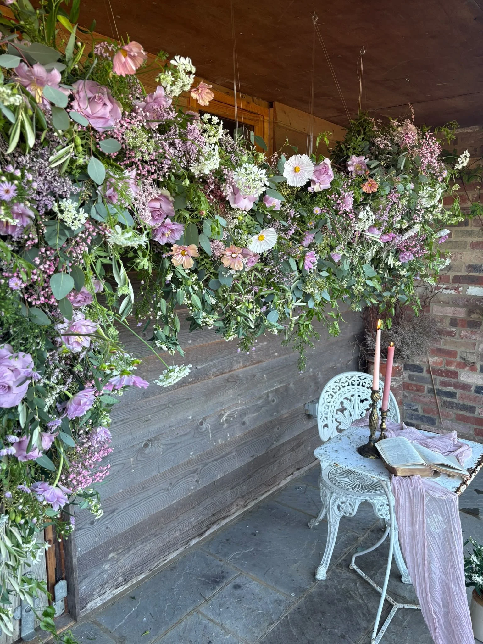 A cozy outdoor space decorated with a large hanging flower arrangement with pink and white flowers and green foliage, a small white ornate round table with a pink cloth, candles, and an open book, against a background of wooden wall panels and a bric