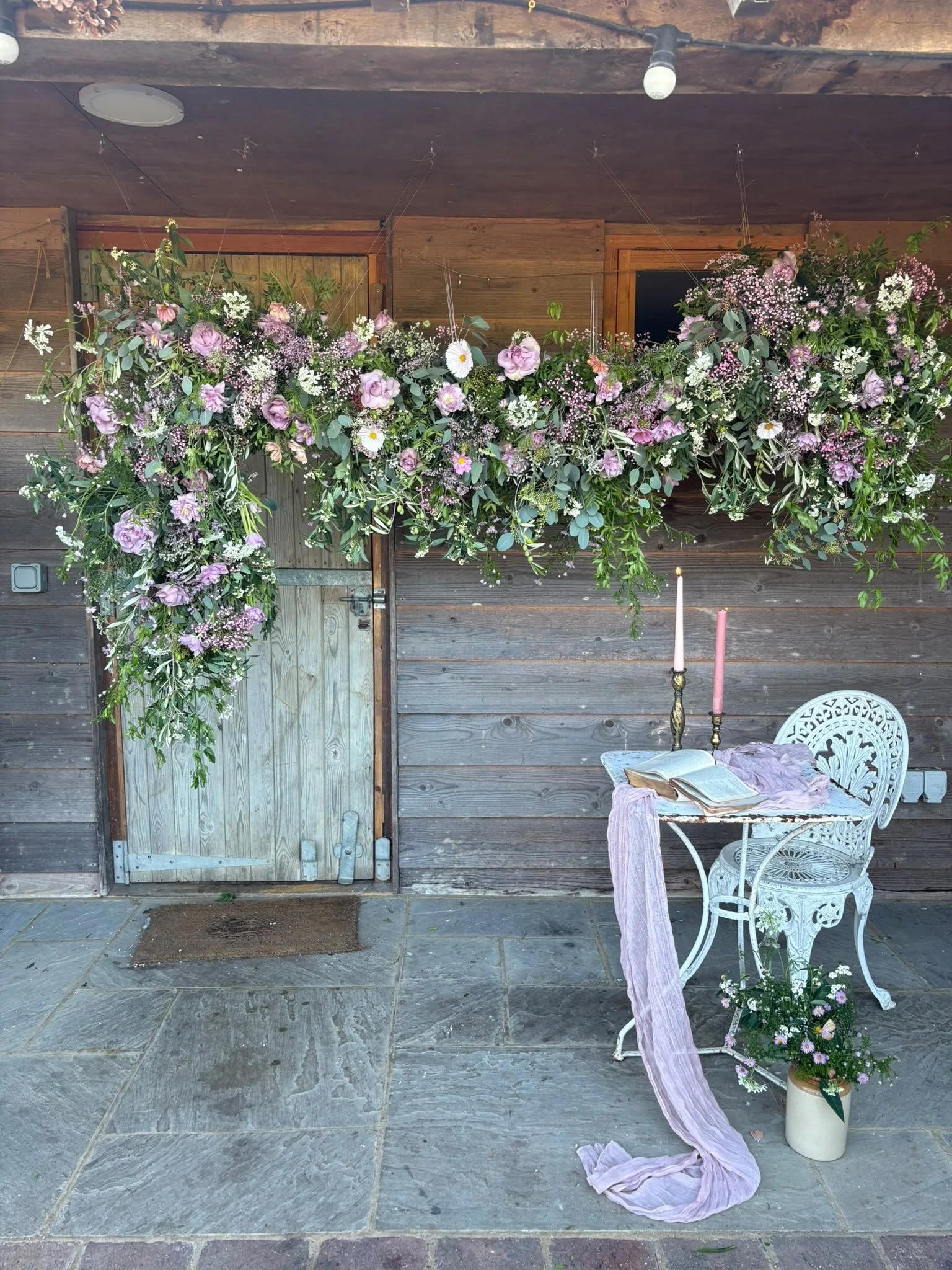 The image shows a rustic wooden wall with a door and two large floral arrangements hanging above. There is a small white metal table and chair set on the stone-tiled floor. On the table, there is an open book, two tall candles in holders, and a pink cloth draped over the table.