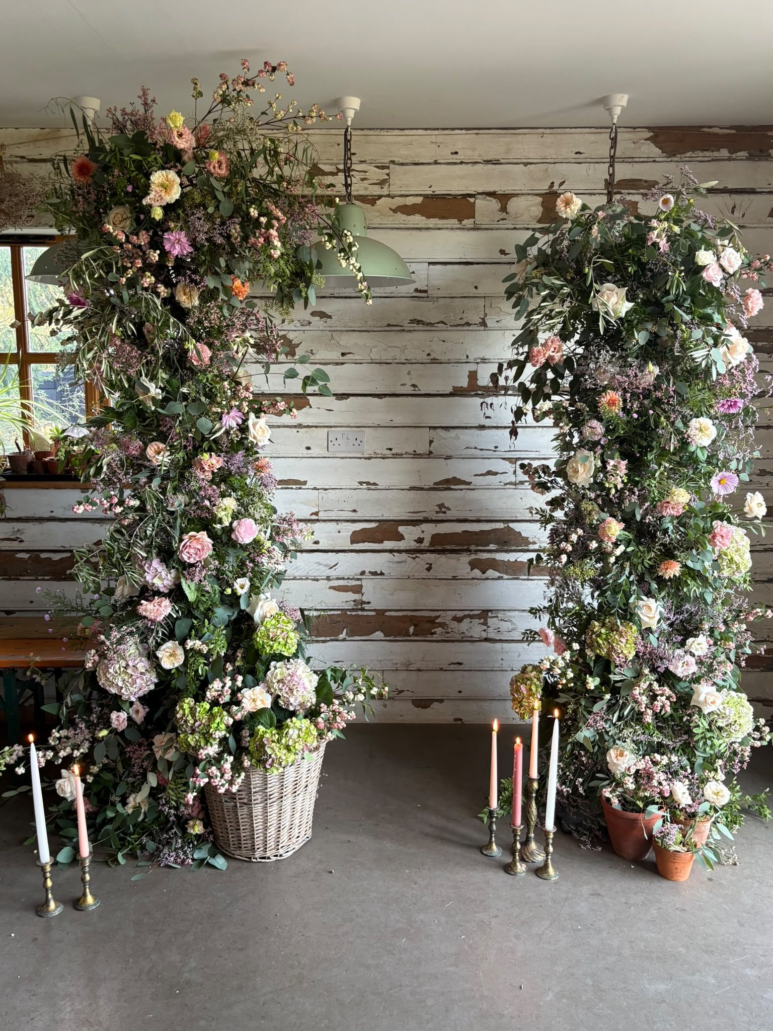 Arrangement of large flower bouquets with pink, white, and purple blooms, placed in white wicker and terracotta pots, set against a rustic white wooden wall with candles in candlesticks in front.